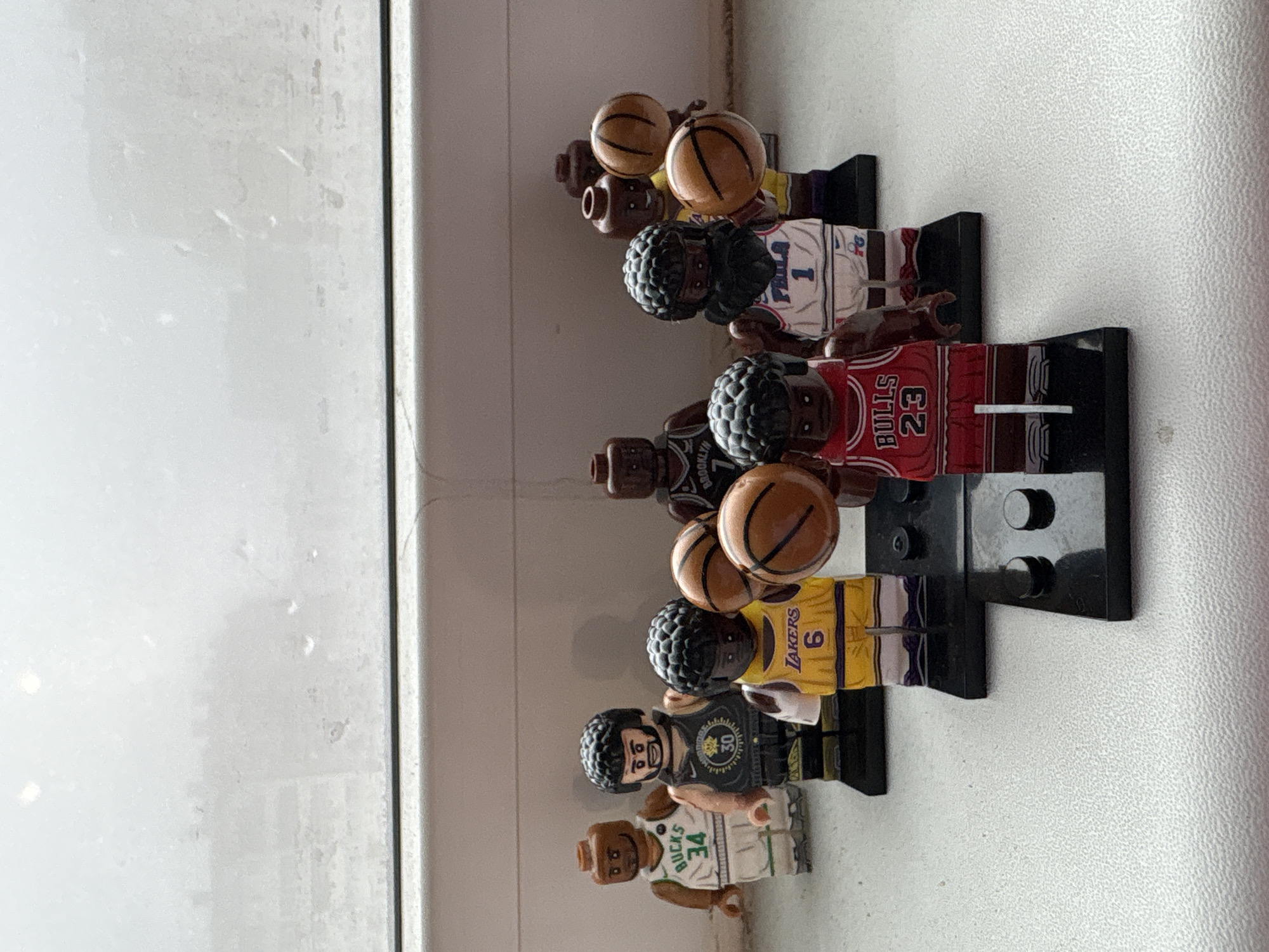 A collection of six LEGO minifigures, stylized as basketball players, stands in a row on a light-colored window sill or ledge in Izhevsk, Russia. The image is oriented vertically, but the minifigures are designed to stand upright. Each minifigure holds a LEGO basketball and is attached to a black LEGO display stand.
From left to right, the minifigures represent:
1. A player wearing a white Milwaukee Bucks jersey with "BUCKS 34" visible, likely Giannis Antetokounmpo.
2. A player in a black Golden State Warriors jersey with "30" on it, likely Stephen Curry.
3. A player in a yellow Los Angeles Lakers jersey with "LAKERS 6" visible, likely LeBron James.
4. A player in a red Chicago Bulls jersey with "BULLS 23" visible, likely Michael Jordan.
5. A player in a dark Brooklyn Nets jersey with "BROOKLYN" and possibly "11" visible, suggesting Kyrie Irving or another Nets player.
6. A player in a white Philadelphia 76ers jersey with "PHILA 1" visible, likely Allen Iverson.
The background consists of a bright, possibly frosted or dirty, window on the left and a light-colored, textured wall on the right. Natural light illuminates the scene, suggesting it is daytime. No specific activity is taking place other than the static display of the figures. The minifigures themselves do not convey emotions.