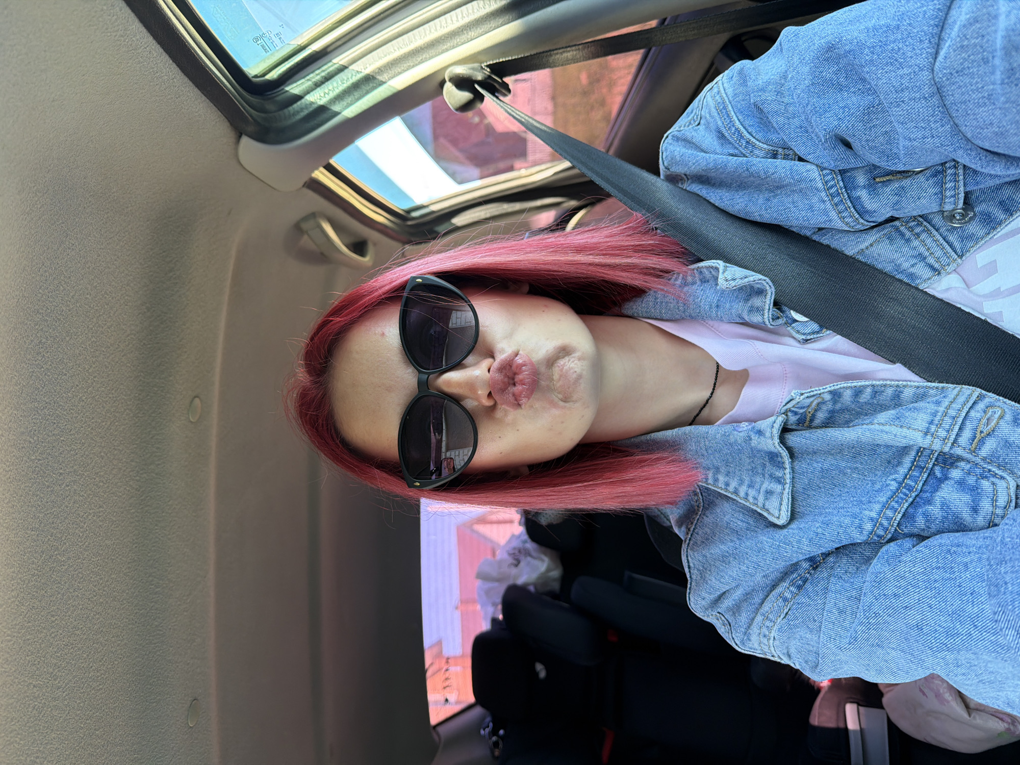A young woman with bright pink hair and sunglasses is seated in the passenger seat of a vehicle, facing forward. She is wearing a denim jacket over a pink t-shirt and a seatbelt is fastened across her body. The interior of the car is visible, including car seats in the background. The scene appears to be a street in Chaikovsky, Russia, with buildings visible through the car windows.