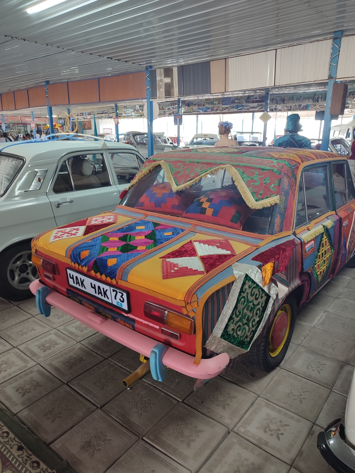 The image shows a highly decorated, vintage car, possibly a Lada, displayed indoors. The car's exterior is covered in a patchwork of colorful textiles, featuring geometric patterns and tassels. The license plate reads "ЧАК ЧАК 73" with a small flag of Kazakhstan below it. The car is parked on a tiled floor, and other vehicles are visible in the background, along with what appears to be displays or exhibits. The ceiling is made of corrugated metal. In the background, two mannequins or figures are visible, one wearing a blue headscarf and the other a floral headdress. The environment suggests a museum or exhibition space in Qonaev, Kazakhstan, dedicated to showcasing vehicles or cultural artifacts.