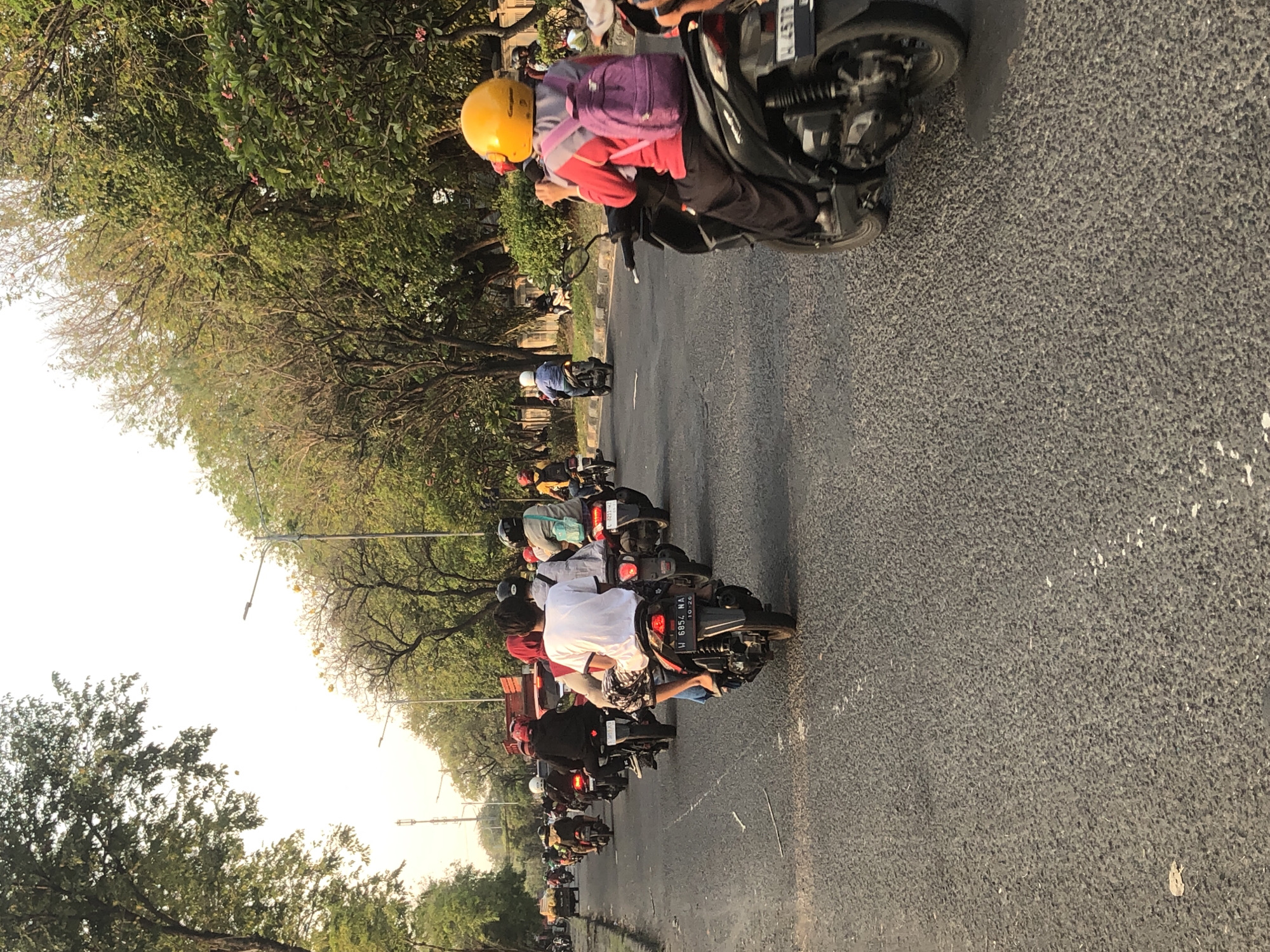 In Gelam, Indonesia, a busy street scene unfolds with multiple motorcycles navigating the asphalt road. The foreground features a motorcycle with a rider wearing a bright yellow helmet and a purple backpack, indicating a journey or commute. Behind them, a procession of other motorcycles fills the lanes, with riders and passengers sporting helmets and various casual attire. The road is flanked by lush green trees and a sidewalk area, suggesting a suburban or urban environment. Streetlights are visible, hinting at the time of day, possibly late afternoon or early evening. The overall atmosphere depicts a common mode of transportation in the region, with a dynamic flow of traffic.