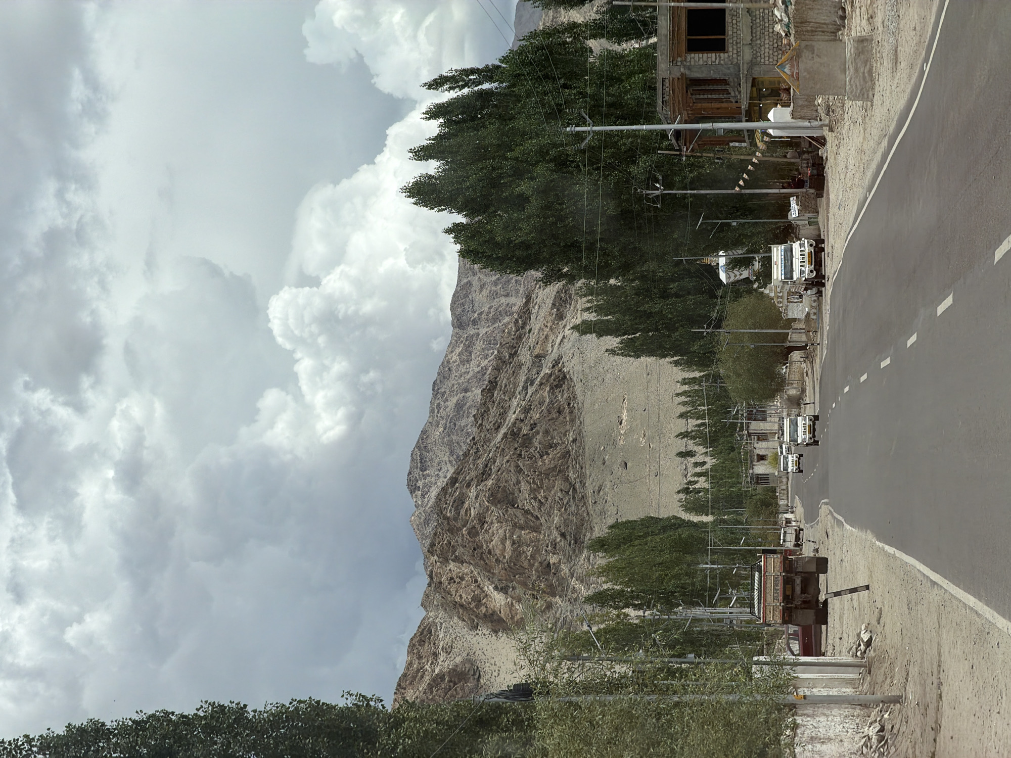This image depicts a view of the road leading into the city of Thiksey, India. In the foreground, a paved asphalt road curves to the right, marked with white dashed lines. On the left side of the road, a dirt shoulder is visible, lined with trees and a white wall. A large, dark-colored truck is parked on the shoulder. A red car is partially visible behind the truck. Power lines and utility poles run along the left side of the road.
Further down the road, several white SUVs are in motion, heading towards the viewer. On the right side of the road, there are buildings with stone and concrete construction. One building appears to be a shop or restaurant with an open facade and awnings. Various signs are visible, including one with colorful prayer flags.
In the background, a range of rugged, rocky mountains dominates the skyline. The sky is filled with dramatic, light-colored clouds, suggesting an overcast or partly cloudy day. The overall scene conveys a sense of a semi-arid, mountainous region.