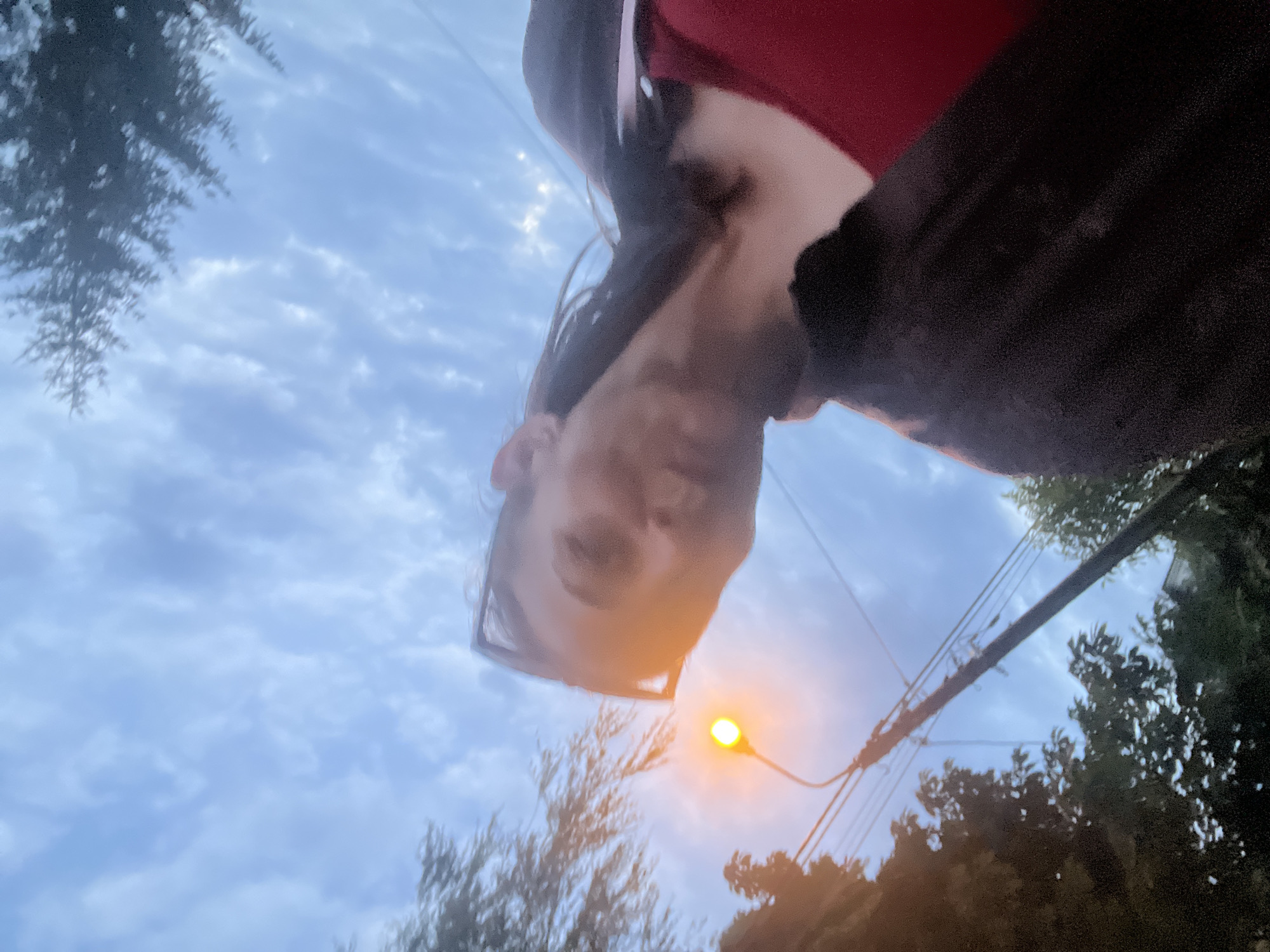 An outdoor, low-angle, eye-level selfie shows a woman from the chest up. The woman has long, dark hair, and is wearing a red tank top, a dark jacket, and sunglasses pushed up on her head. She is looking at the camera. Behind her, a bright orange street lamp glows against a cloudy, blue sky. Trees and power lines are visible in the background. The image is slightly blurry and overexposed due to the street lamp.