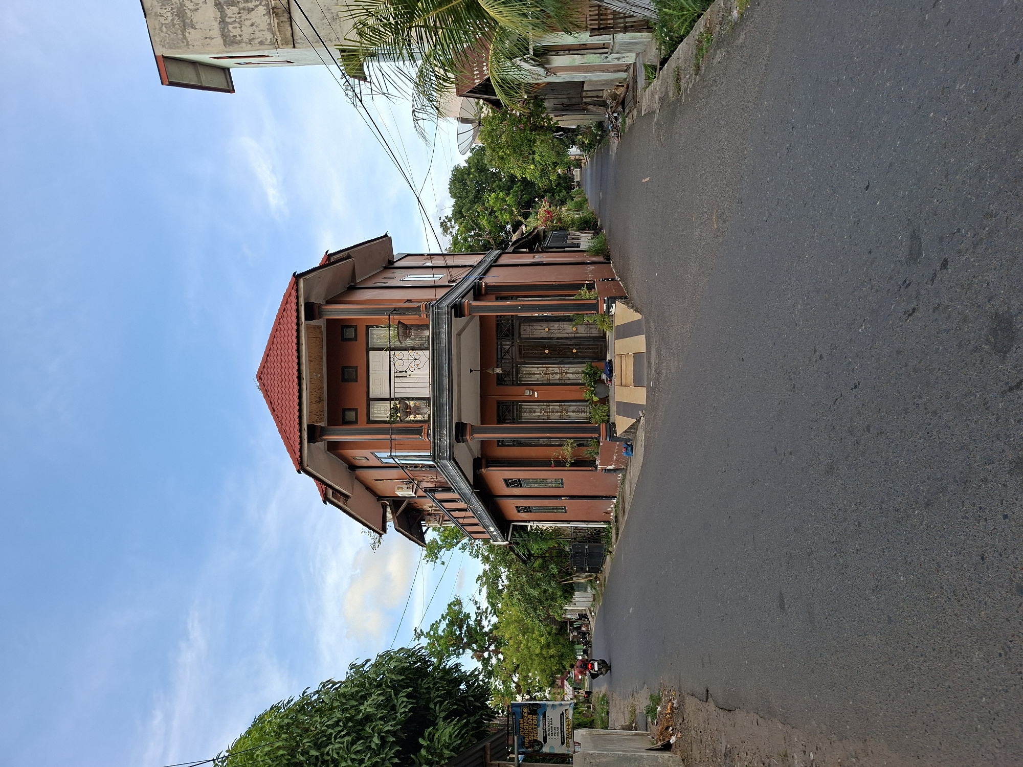 The image depicts a street scene in Lhokseumawe, Indonesia. A two-story building with a red tile roof and a brown facade dominates the left side of the frame. The building features a balcony with ornate metalwork and multiple windows and doors. The street is paved with asphalt and appears relatively narrow. Power lines stretch across the sky above the buildings. On the right side of the street, vegetation, including trees and foliage, lines the road. In the distance, the street curves and appears to lead into a more densely vegetated area. A small motorcycle is parked on the side of the street near the bottom of the frame. A sign is partially visible near the motorcycle. The sky is blue with scattered clouds.