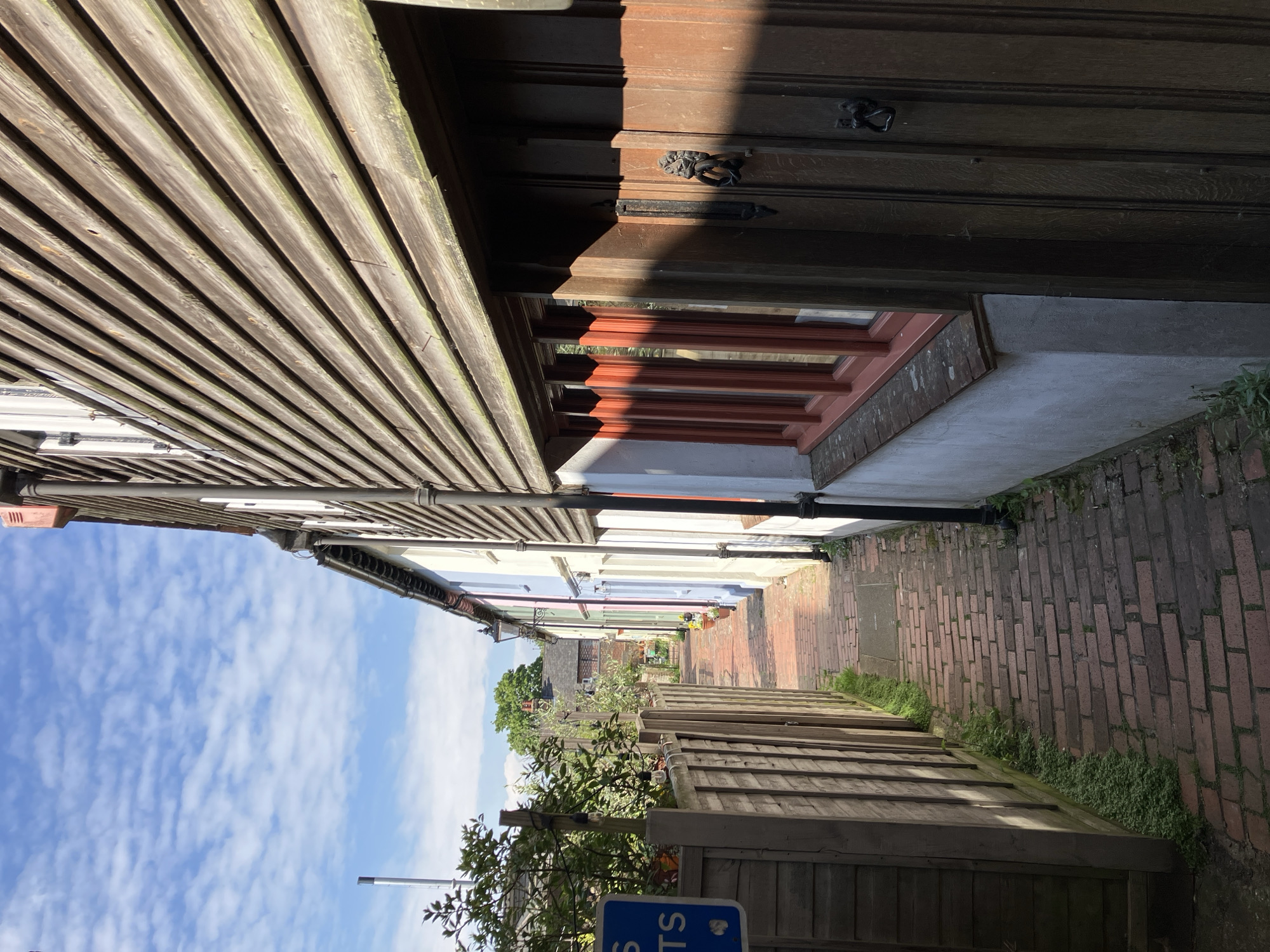 The image presents a narrow alleyway in Lewes, United Kingdom, paved with red bricks. Wooden fences and building façades border the path on either side. The buildings exhibit various colors, including white, blue, and pink. At the end of the alley is green foliage. The sky above is blue with scattered white clouds. A wooden structure looms on the left, with horizontal planks. A wooden door is to the right, adorned with metal fittings and a window below. A black pipe runs along the wall. A blue sign is visible near the bottom of the image.