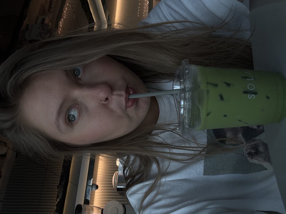 A young woman with blonde hair and blue eyes is holding a green drink. She has a nose ring and is drinking from the cup with a straw. The drink is a light green color with ice cubes. The brand name "SOLA" is printed on the cup. The woman is wearing a white t-shirt. The background is blurred, but it appears to be a cafe or restaurant. The picture is angled.