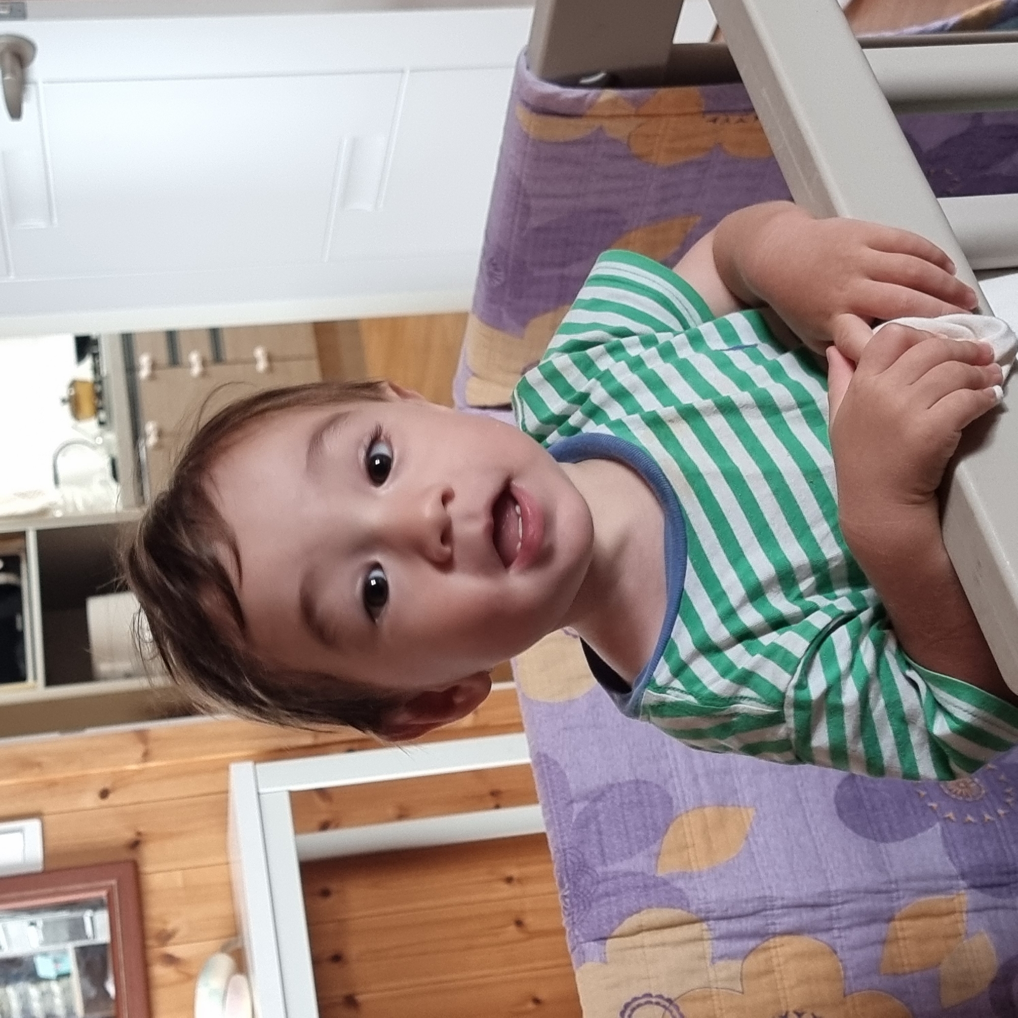 A young child is positioned in the foreground, looking directly at the camera. The child is wearing a green and white striped t-shirt with a blue trim around the neckline. Their hands are resting on a light-colored railing in front of them, and they are holding a white cloth. Behind the child, a blanket with a purple and yellow floral pattern is visible, draped over a railing. The background features wooden paneling on the left and a white door on the right, which has two rectangular indentations and a silver handle. Further in the background, a kitchen counter with appliances and cabinets can be seen. The scene appears to be indoors.
