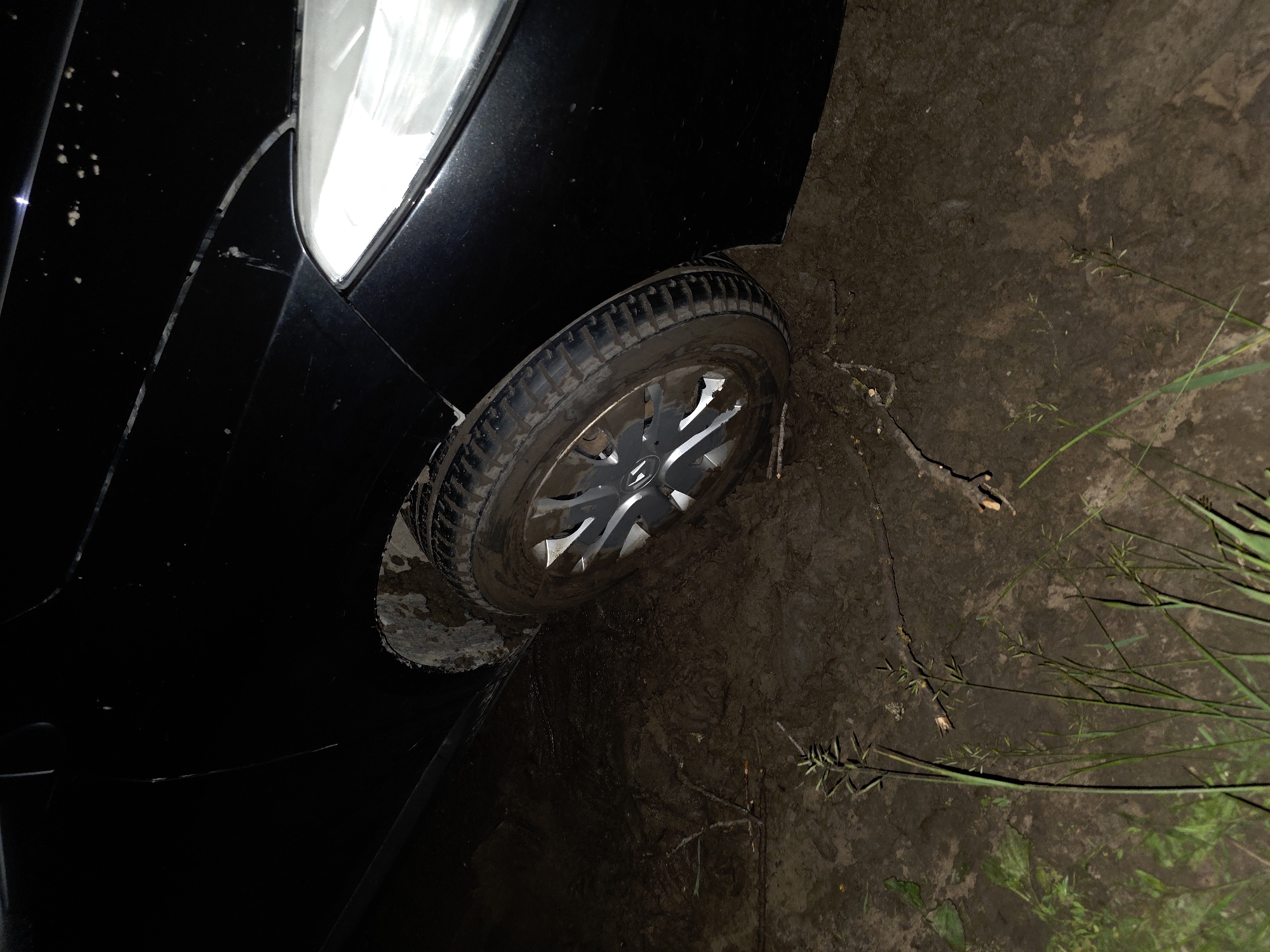 The image shows a black car stuck in mud. The front passenger-side wheel of the car is partially submerged in thick, wet mud. The car's fender and headlight are visible, as is the tire and rim of the stuck wheel. The surrounding environment is muddy ground with some sparse vegetation and twigs scattered on the surface. The scene appears to be outdoors, possibly in a rural or undeveloped area, with no clear signs of city infrastructure. The lighting suggests it may be nighttime or a very dimly lit area. The specific location in the city of Tagashevo, Russia, is not discernible from the visual information in the image alone.