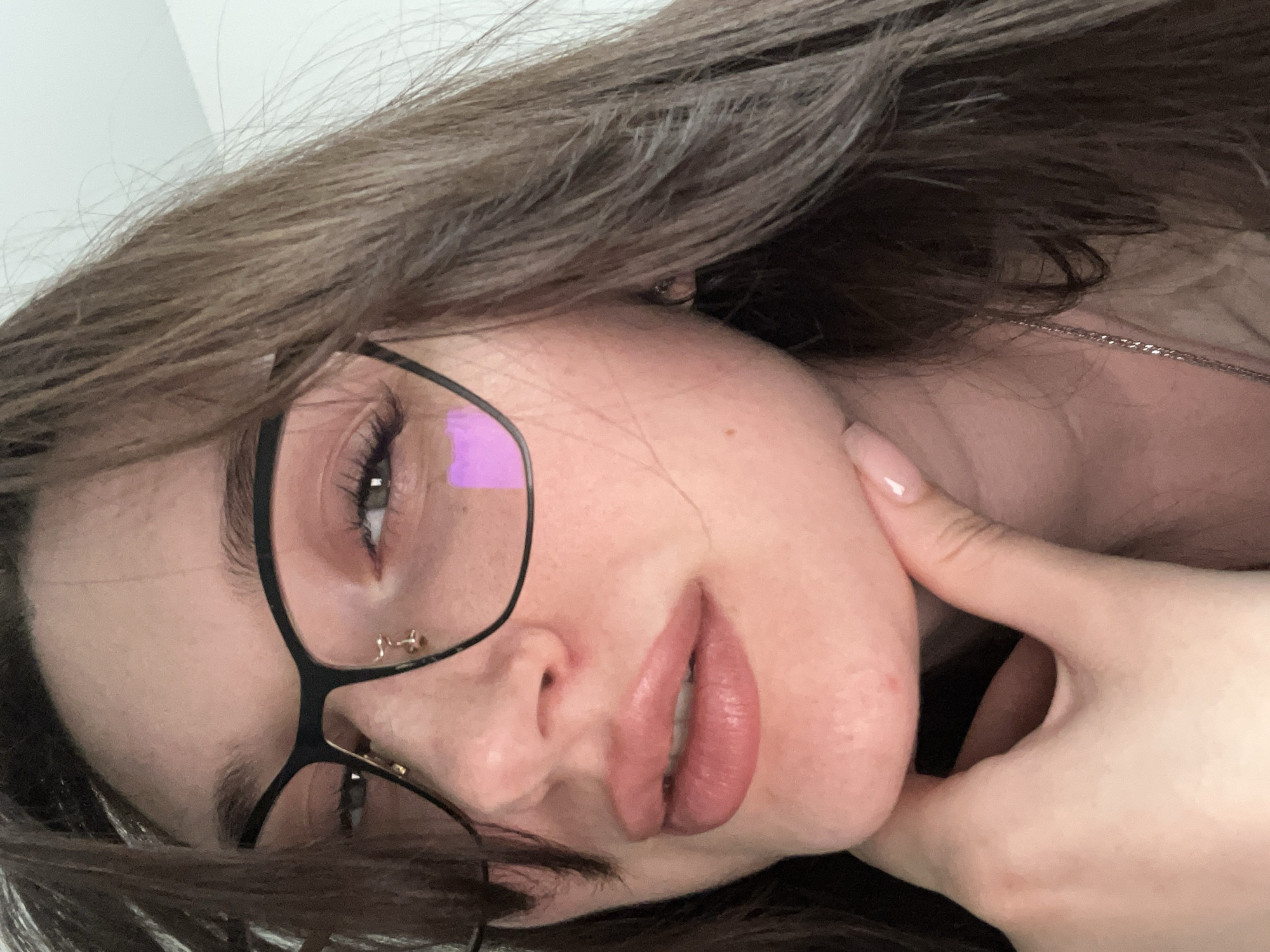 A close-up, rotated portrait depicts a young woman, likely a selfie taken indoors. Her head is tilted to her right, with her long, brown hair partially covering her shoulders and face. She wears black-framed glasses, reflecting a distinct purple hue, possibly from a screen or light source. Her eye is visible behind the lens, accentuated by long eyelashes. A small, golden piercing is visible near her eye. Her lips are full, colored with a light pink or nude lipstick. Her left hand, adorned with light pink manicured nails, gently touches her chin and jawline. A thin silver chain necklace is visible around her neck. The background is out of focus, appearing as a plain, light-colored surface. The woman's expression is neutral as she looks towards the camera. There is no visible text in the image. The location context provided is Tyumen, Russia.