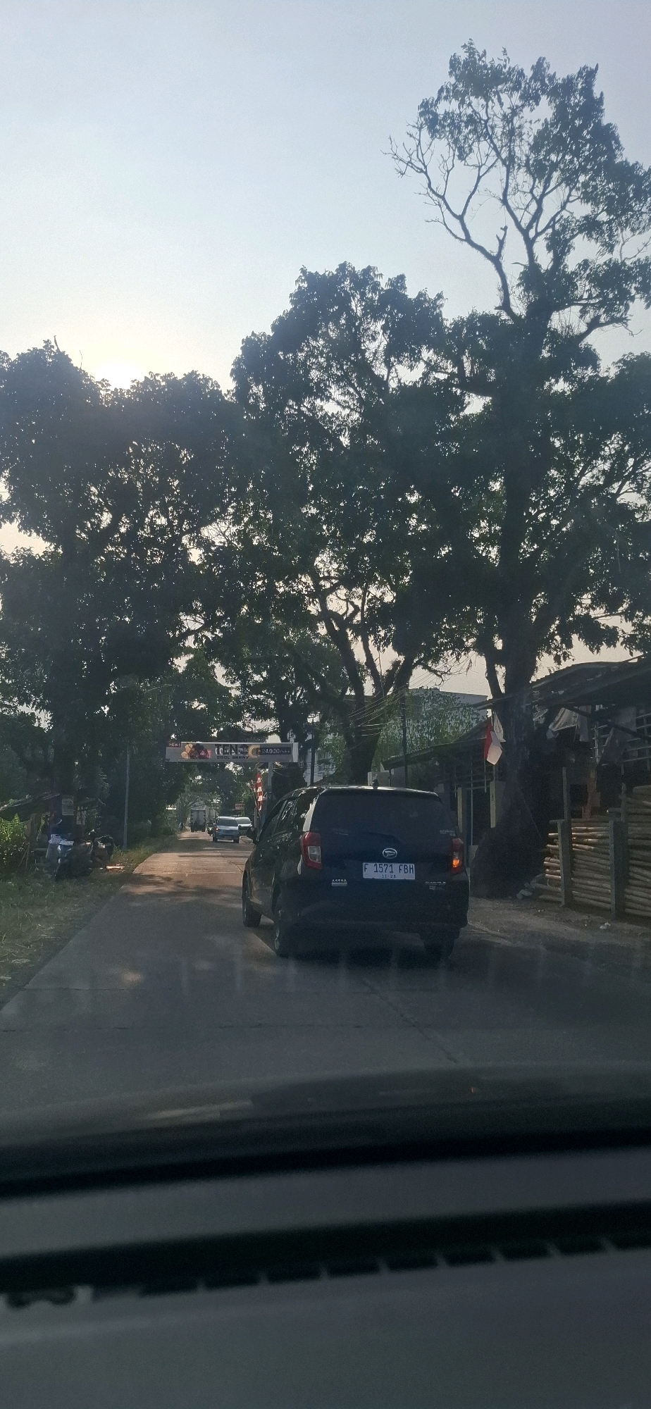 The image shows a street scene in Sukasari, Indonesia, with a black Daihatsu hatchback in the foreground, its rear facing the camera. The car has a license plate that reads "F 1571 FBH". The vehicle is driving on a paved road lined with trees. Overhead, a large banner displays the text "TERIMA KASIH TELAH BERKUNJUNG". On the right side of the road, there are some buildings and fences made of bamboo. The sky is overcast with the sun setting, casting a soft light over the scene. There are other vehicles on the road in the distance. The image is taken from inside another vehicle, looking forward.