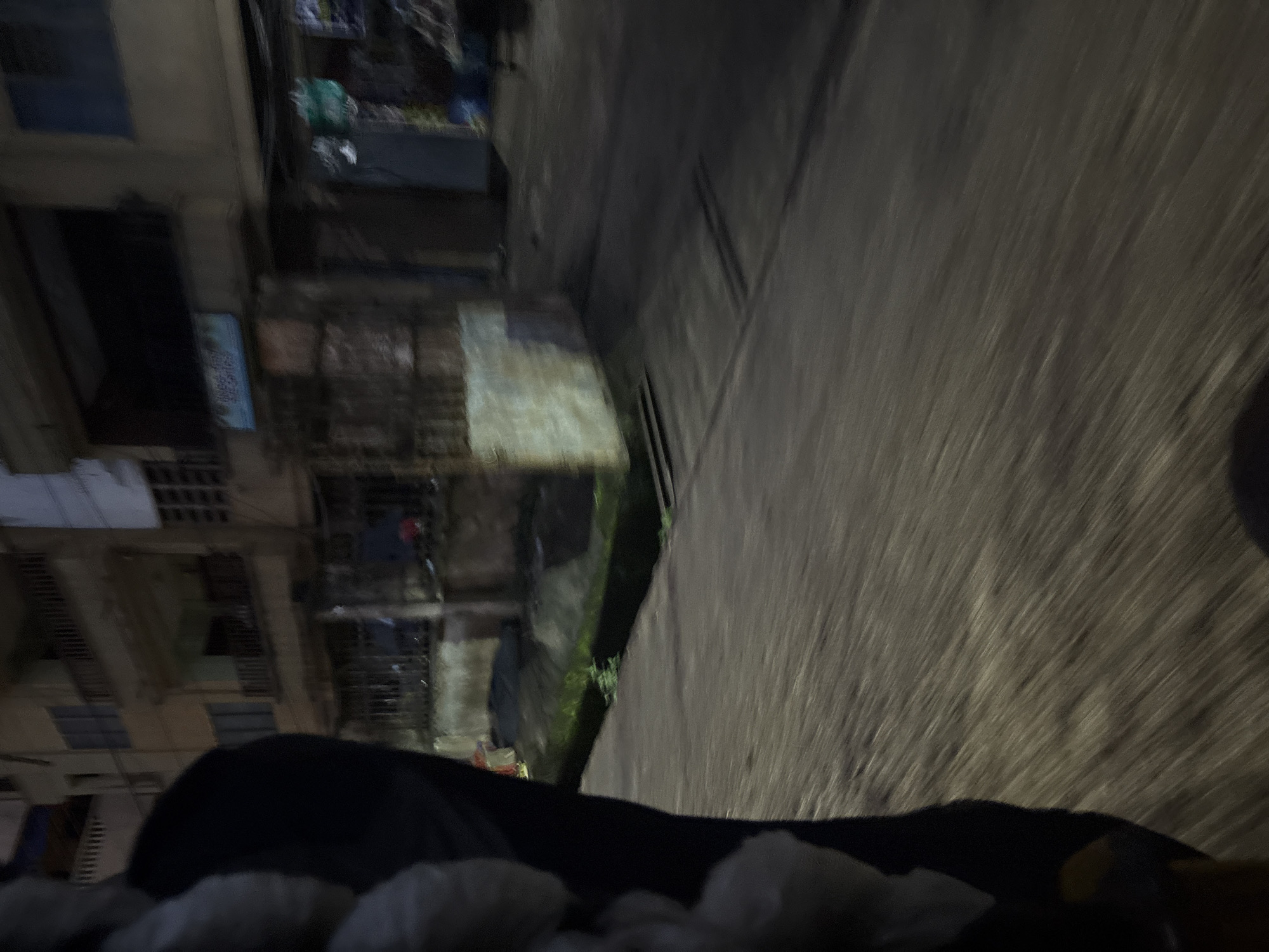 This is a low-light, motion-blurred image taken from a vehicle moving on a paved road in Onitsha, Nigeria. In the foreground, the dark silhouette of a person's shoulder and arm is visible on the left, with what appears to be white fabric or packaging. The road surface is gray and textured, with streaks indicating movement. To the right and in the background, a multi-story building with balconies and windows is partially visible. The building appears to be in disrepair, with some areas dark and others illuminated by faint light. A blue sign with text in an Asian script is affixed to the building. There is a narrow drainage ditch with some vegetation running alongside the road in front of the building. The overall impression is a snapshot of urban life at night in Onitsha, characterized by movement and low visibility.
