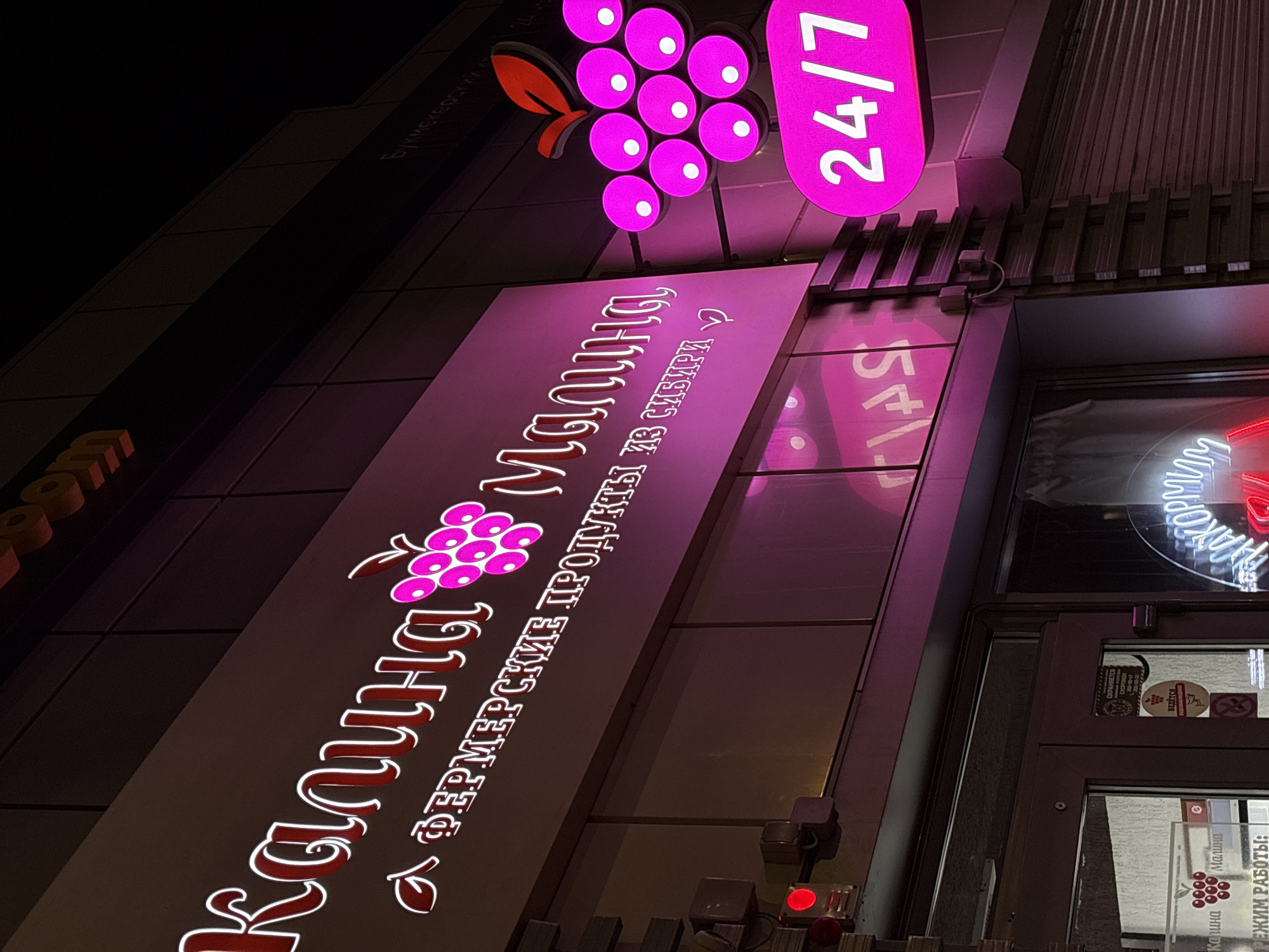Captured at night, the image features the storefront of "Kalina Malina," a Siberian farmer's market, located in Krasnoyarsk, Russia. The shop's signage, illuminated in red and magenta, displays its name in Russian alongside a grape cluster design. Further signage indicates the store is open 24/7. Below, a door is visible, displaying stickers and information about the business hours. The surrounding environment appears dimly lit, suggesting an urban setting at night.