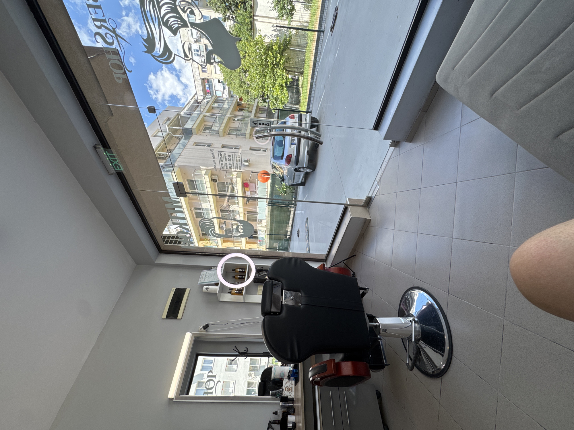 The image depicts the interior of a barbershop in Plovdiv, Bulgaria, viewed from the perspective of a seated individual. The foreground features a section of a person's leg and a portion of a gray upholstered bench. The middle ground is dominated by a black barber chair with red accents, positioned on a metallic base. To the left of the chair, a gray counter with drawers holds various grooming products. A mirror with an illuminated frame hangs on the wall above the counter. A shelf unit to the right of the chair displays several bottles of hair products.
The background reveals the exterior through a large glass entrance. A silver car is parked on the street. Buildings with balconies and an urban sky with clouds are visible. A sign with the word "EXIT" is illuminated above the glass door. The floor inside the barbershop is tiled, and the walls are painted gray. The overall impression is a modern and clean barbershop environment.