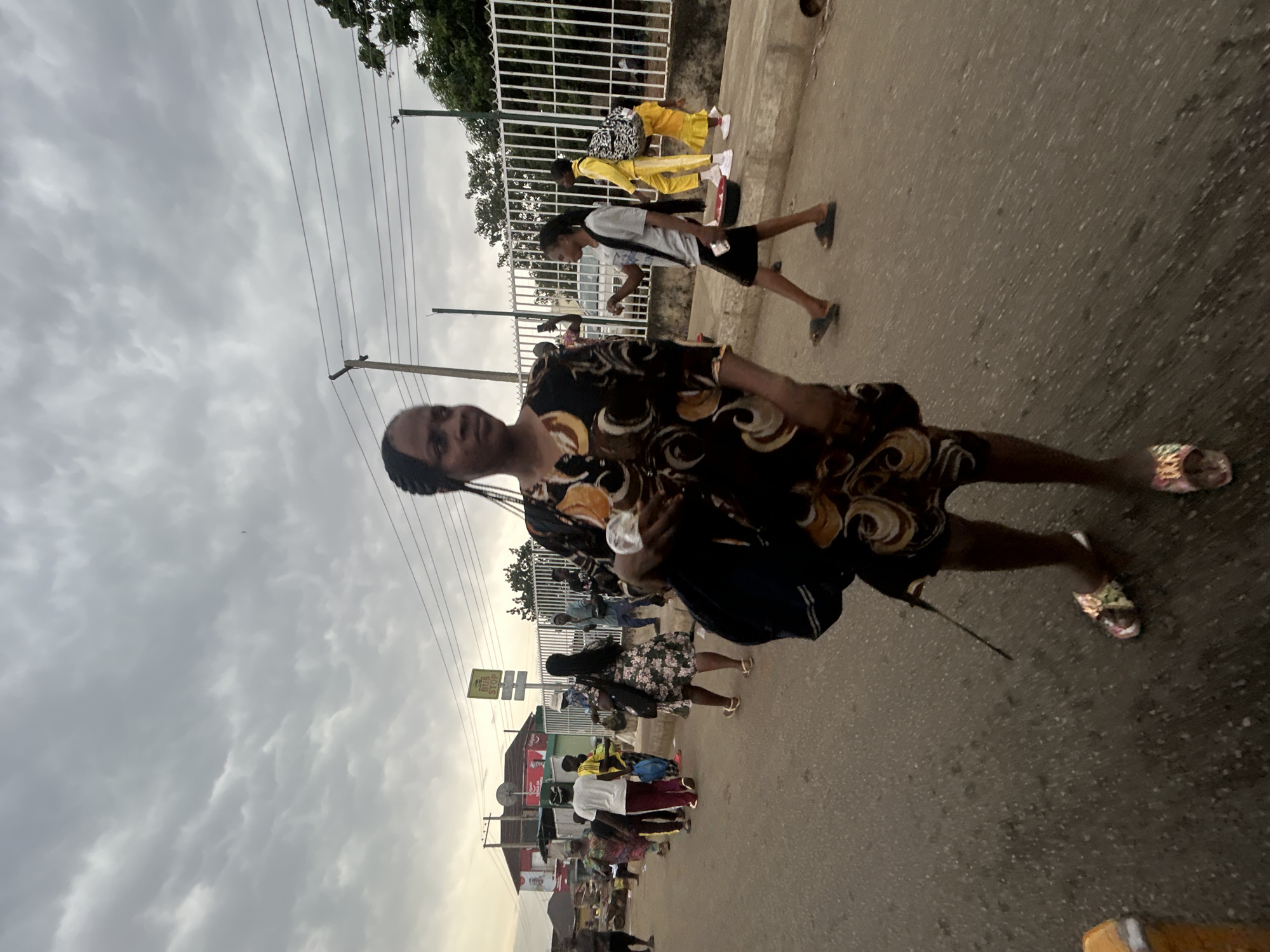 The image depicts a street scene, possibly a commercial area, in Jos, Nigeria, under an overcast sky. A woman wearing a dark dress with abstract patterns is walking towards the camera on the right side of the frame. Several other individuals are walking along the street, including a person in a yellow tracksuit and another in a white shirt and dark shorts. A bus stop sign is visible along with other buildings and shops. Overhead power lines stretch across the sky. The road surface is asphalt.