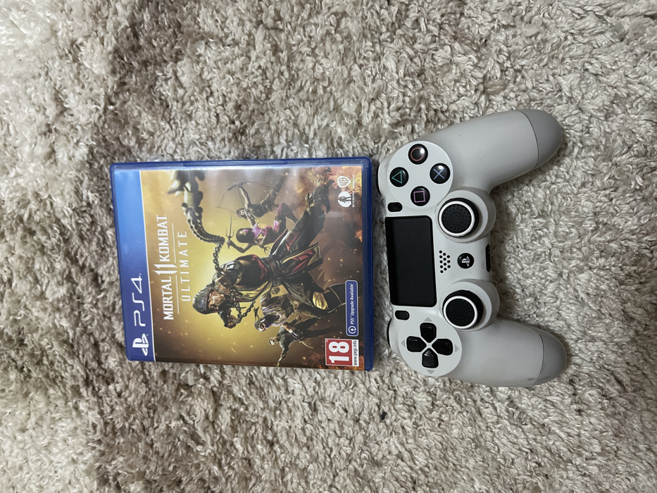 An overhead view depicts a light grey PlayStation DualShock 4 controller positioned directly in front of a blue PlayStation 4 game case for "Mortal Kombat 11 Ultimate." The game case features key art of multiple characters, with Scorpion prominently centered, displaying chains. Visible text on the case includes "PS4," "MORTAL KOMBAT 11 ULTIMATE," an "18" PEGI rating, and "PS5™ Upgrade Available." The controller exhibits its standard button layout, including the directional pad, face buttons (triangle, circle, cross, square), dual analog sticks with black caps, a central touchpad, and the PlayStation logo. Both items rest on a textured, light brown shag carpet. The scene is located in Sedlartsi, Bulgaria.