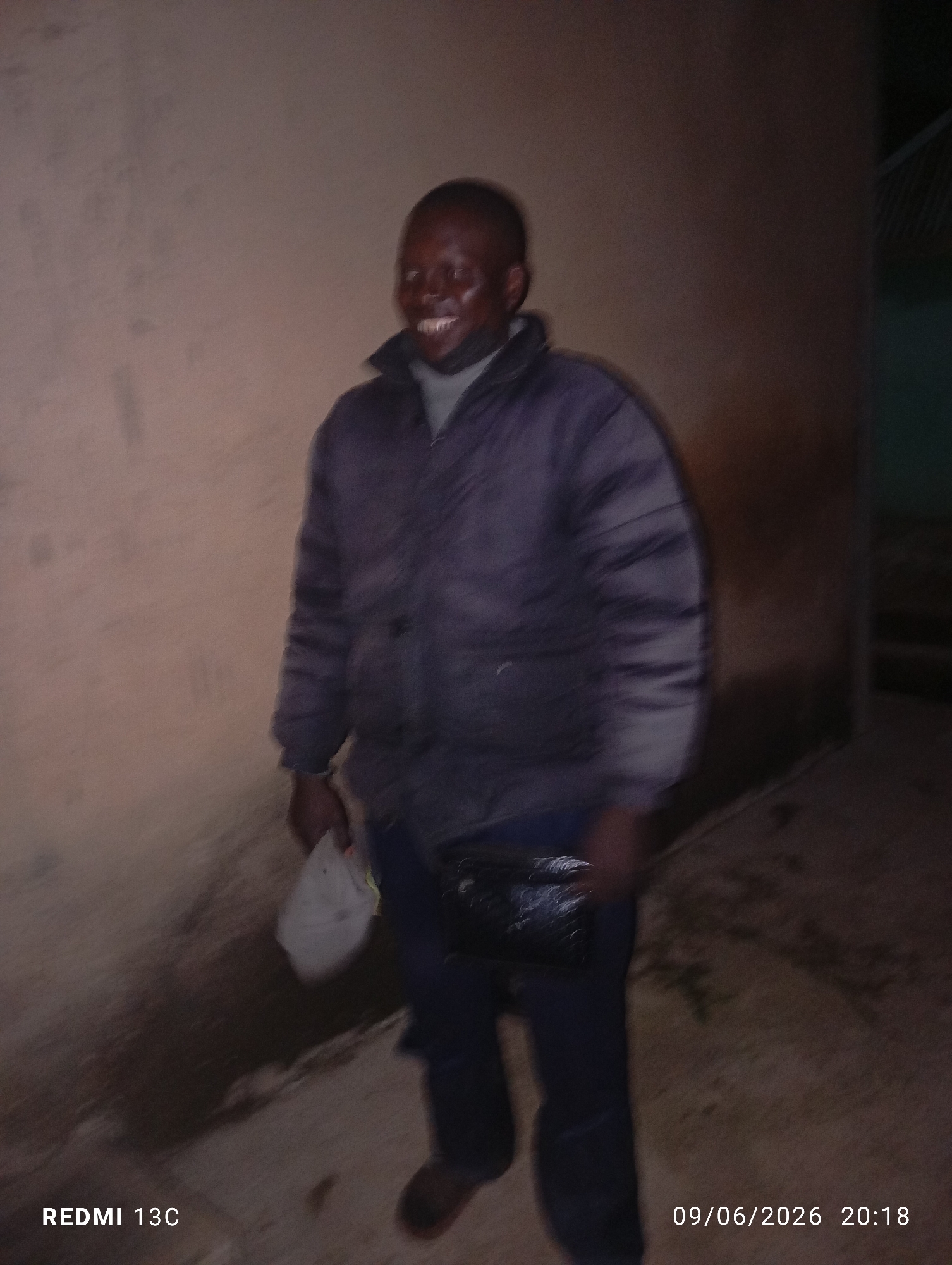 A man in a dark jacket stands outdoors at night. He is smiling broadly. He is holding a white cap in his left hand and a black clutch bag in his right hand. The background is a light-colored wall. A date and time stamp in the lower right corner reads "09/06/2026 20:18".
