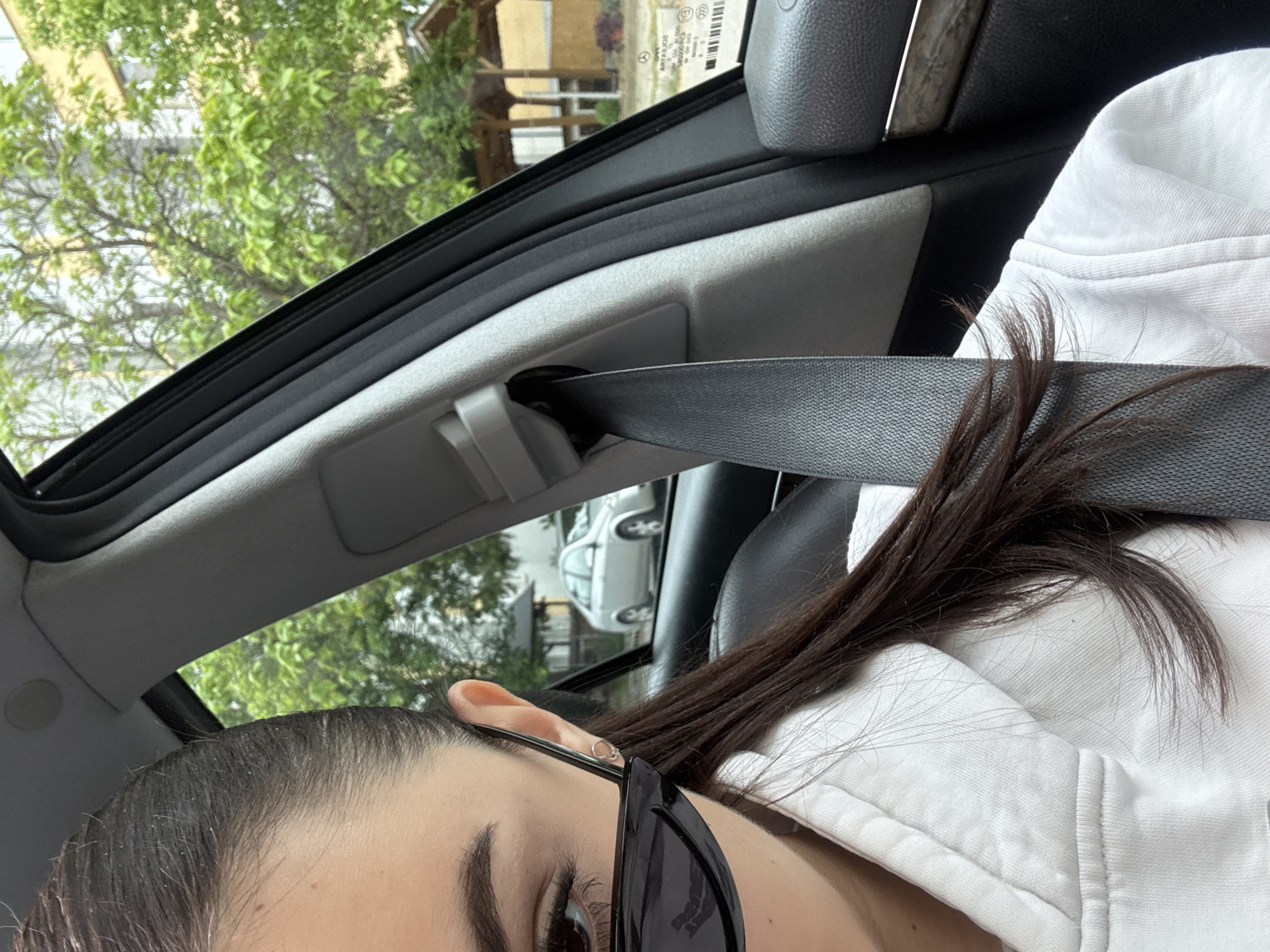 A partially obscured female subject is visible inside a car. The subject is wearing a white hoodie and sunglasses, with a seatbelt across their chest. The car's interior is visible, including the roof and a partially visible car seat. The background shows a street with a building and trees. The exact location is unknown.
