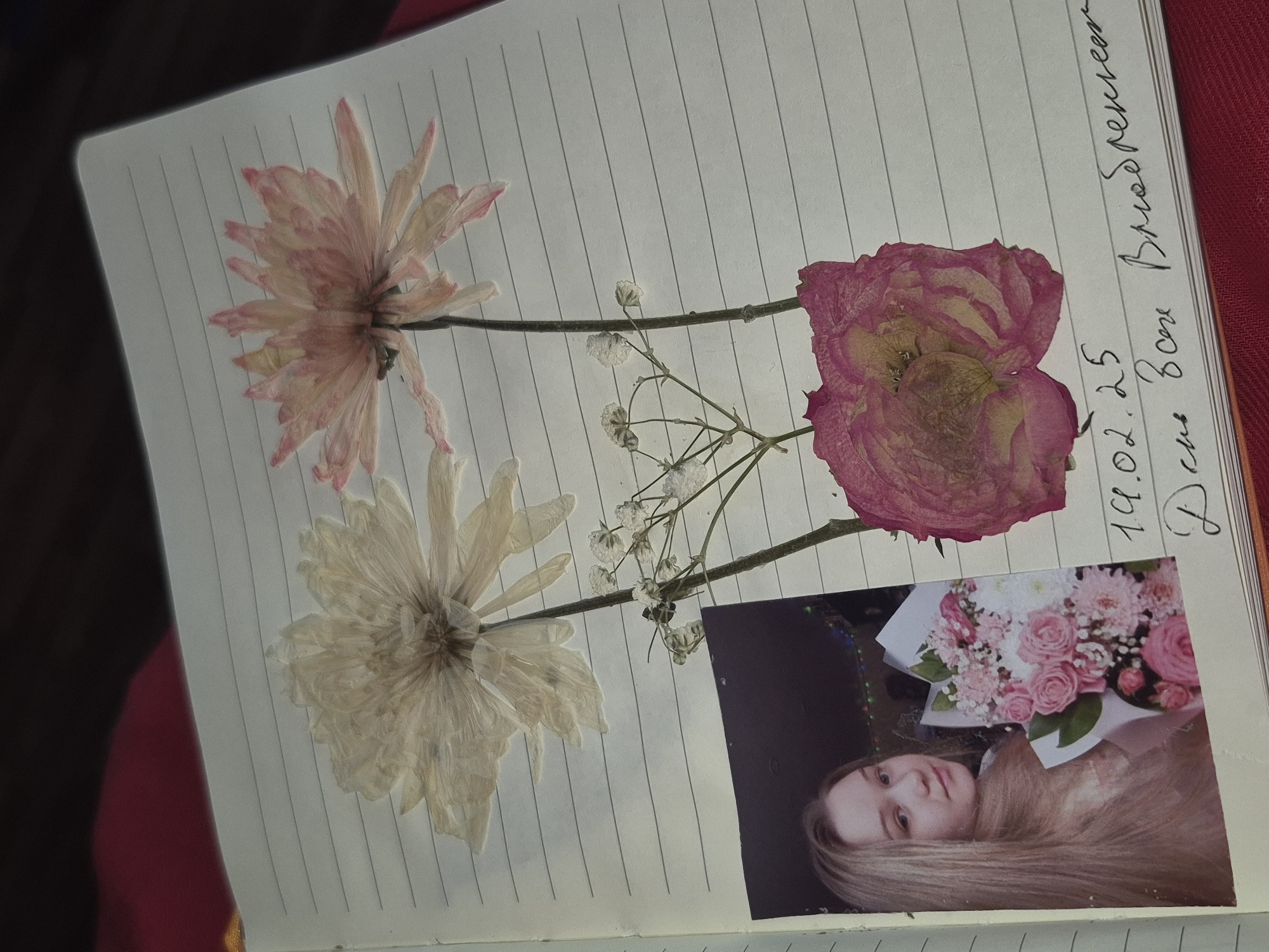 An open notebook lies on a surface, with dried flowers pressed onto its lined pages. The flowers include two daisies, one pink and one white, and a pink rose. A small photograph is affixed to the lower right corner of the page. The photograph depicts a woman with long blonde hair holding a bouquet of pink and white flowers. To the right of the flowers, handwriting indicates a date "19.02.25" and the words "День Всех Влюбленных" (Day of All Lovers, i.e., Valentine's Day). The notebook is assumed to be located in Chaikovsky, Russia based on the language of the handwritten note.