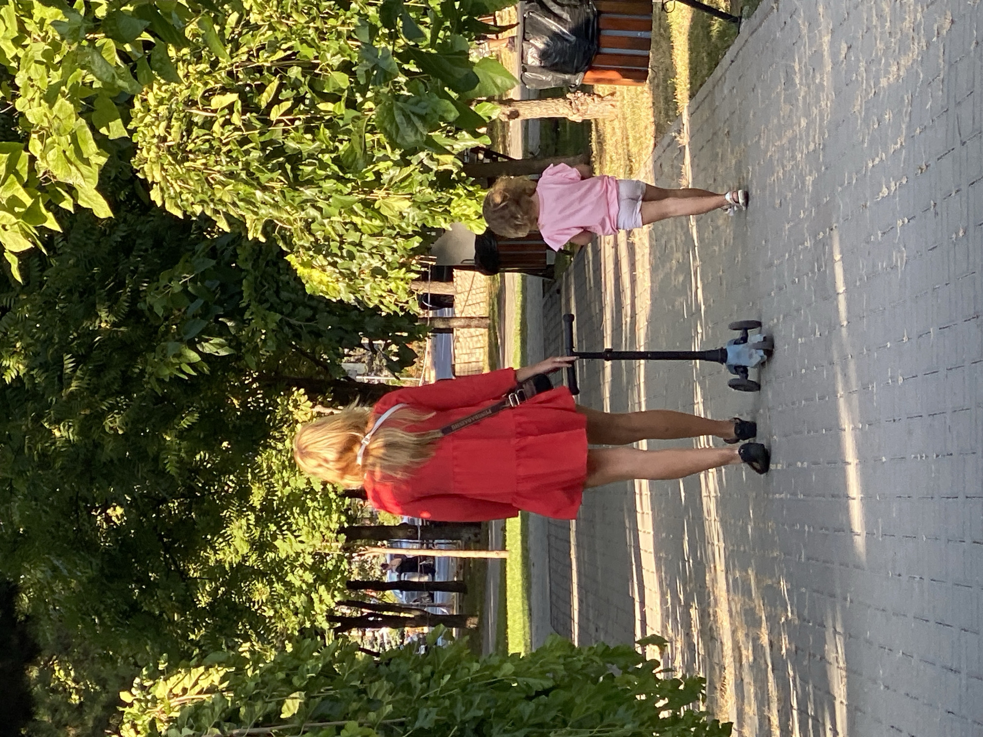 In a sunlit park setting, likely within Chișinău, Moldova, a woman and a young child are walking on a paved pathway. The woman, with long blonde hair, wears a short red dress and a crossbody bag. She is walking towards the viewer's left, partially obscuring a child's scooter. The child, seen from behind, has curly blonde hair and wears a pink t-shirt and light-colored shorts. The child is walking away from the viewer, towards the right. The pathway is bordered by green grass and mature trees with dense foliage, casting dappled shadows on the ground. In the background, trees, some parked cars, and what appear to be park benches and trash receptacles are visible. The overall scene suggests a pleasant day in an urban park.