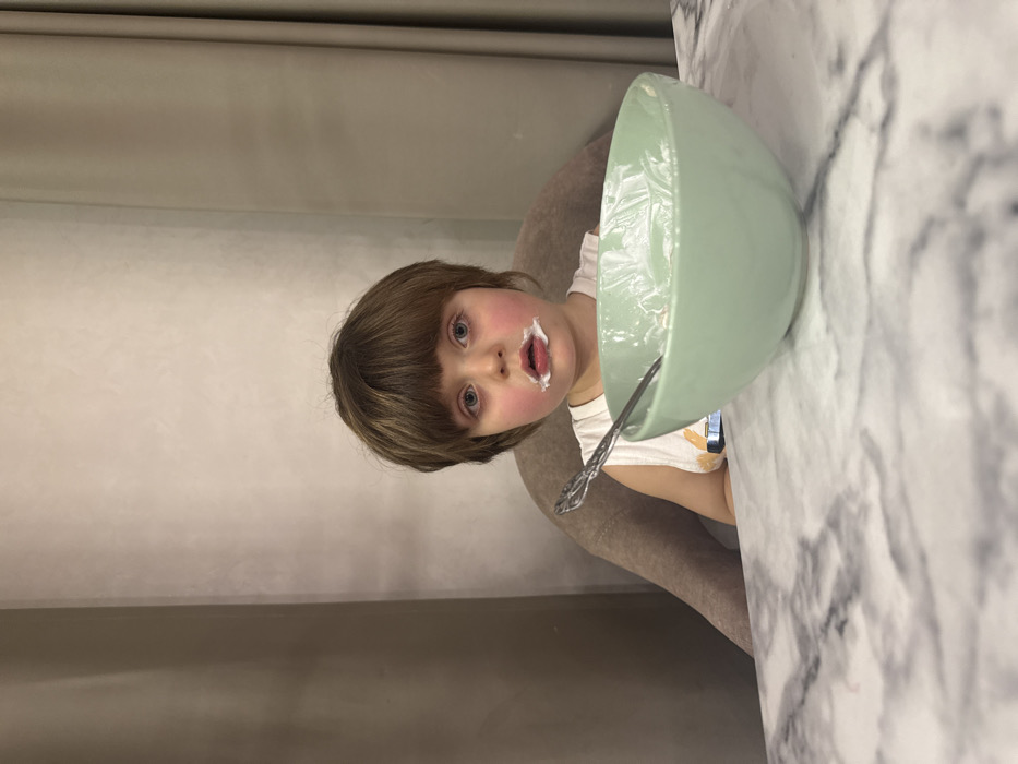A young child with brown hair and blue eyes looks directly at the camera. The child's mouth is open, and there is white food residue around the mouth. In front of the child is a large, light green bowl with a spoon inside. The bowl appears to contain a similar white food. The child sits at a table with a marble-like surface. A neutral-toned chair is visible beneath the child. The background consists of a textured, light brown wall.