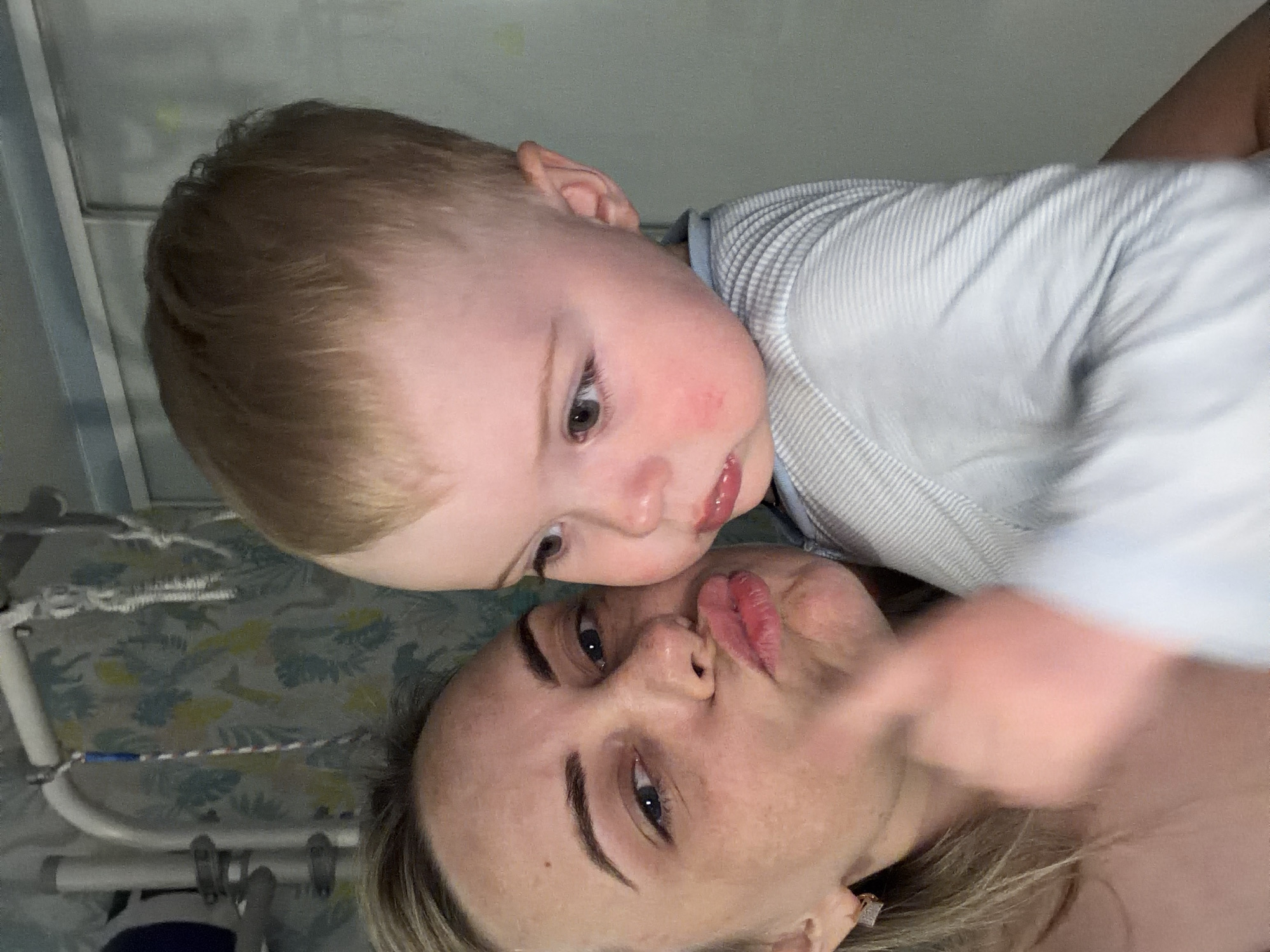 A young Caucasian child is lying on top of a light-skinned adult. The child has fair hair and is wearing a light-colored striped shirt. The adult has blonde hair and is wearing an earring. The background includes part of a hospital bed with a patterned sheet and a railing. The location is possibly a hospital room, potentially in the city of Votkinsk, Russia.