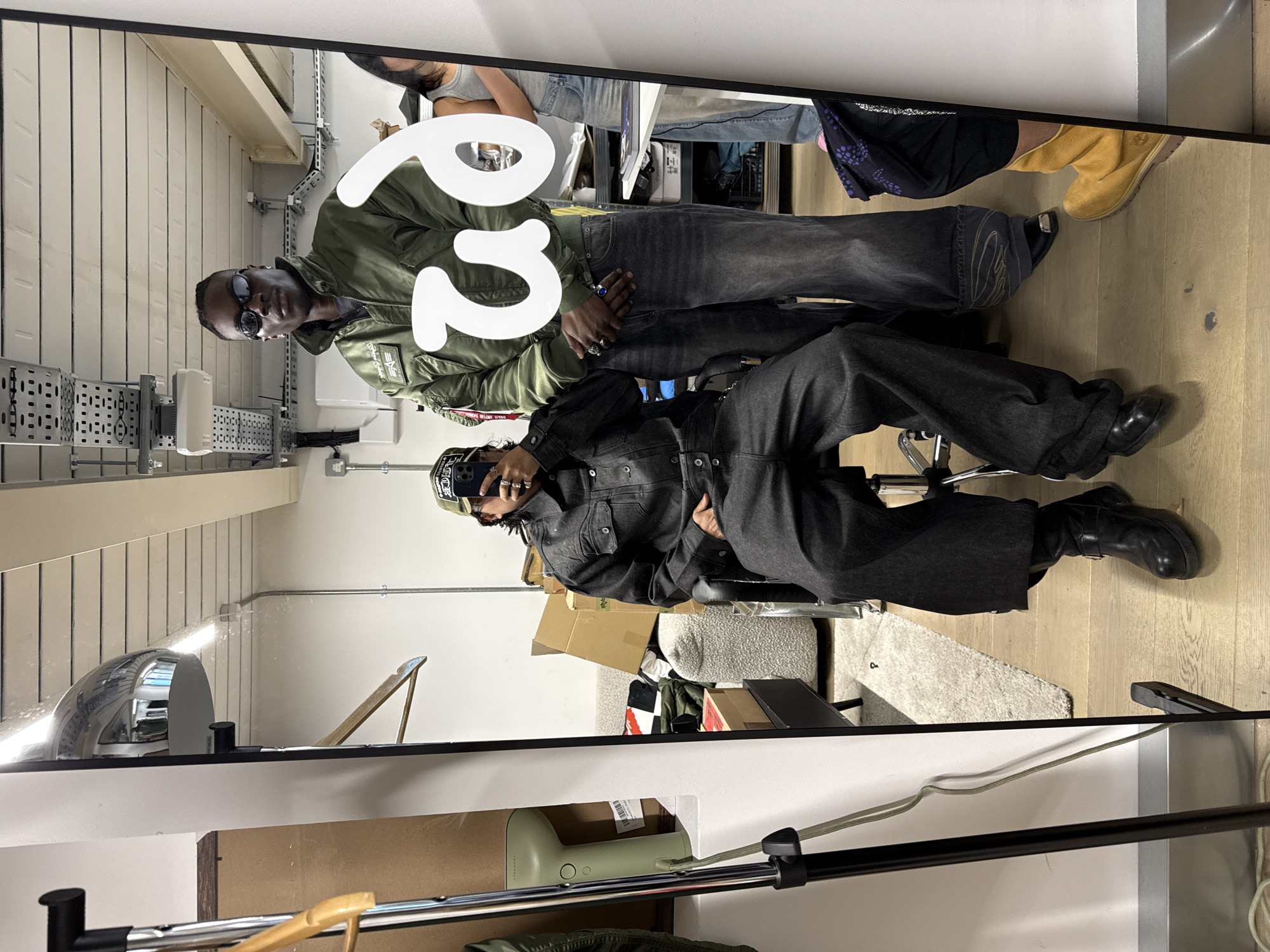 This indoor mirror selfie captures two individuals and their surroundings, presented rotated 90 degrees counter-clockwise but described as if upright. The setting appears to be a modern studio or workspace in London, characterized by light-colored wooden floors, white walls, and industrial-style metal cable trays and conduits running along them.
In the foreground, a person with dark skin is seated on a black office chair, dressed in a dark denim jacket and matching wide-leg pants, paired with black boots. They are holding a smartphone (likely an iPhone) to take the photograph, with rings visible on their fingers and a cap covering their head. Their face is largely obscured by the phone and cap.
Standing behind and slightly to the left of the seated person is a Black man wearing dark sunglasses, an olive green bomber jacket (with an "Alpha Industries" patch and a red "Remove Before Flight" tag on the sleeve), and dark jeans. His hands are clasped in front of him, and he looks directly into the mirror.
Large, abstract white shapes or numbers, resembling a "2" and part of a "9" or "6", are overlaid in the center of the image, obscuring parts of the standing man's torso.
The background reflection shows more of the room: cardboard boxes, a white fluffy rug, and glimpses of other people, including a person in a grey tank top and light-wash jeans, and another person's leg wearing a tan boot. Visible objects also include a chrome-domed light fixture, a light green appliance (possibly a humidifier, with "STRAWBERRY" and "MADE IN CHINA" on its packaging), and a wooden hanger. The overall activity is a casual mirror photo shoot in an urban, functional space.