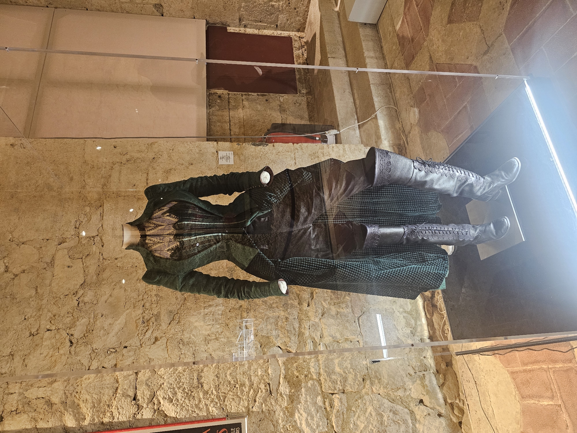 The image shows a display of a historical costume, likely from a film or theatrical production, in a glass-enclosed exhibit. The costume consists of a dark green, high-necked jacket with puffed shoulders and intricate detailing, worn over a black corset with lace trim. A dark, textured skirt with a diamond pattern is visible beneath the jacket, and the ensemble is completed with tall, black leather lace-up boots.
The exhibit is set against a rustic stone wall, typical of historical buildings. Behind the costume, a fire extinguisher is visible on the wall, and beyond that, a set of stone stairs leads to a dark opening, possibly a doorway. The floor appears to be paved with terracotta tiles. A sign is attached to the wall near the costume, likely providing information about the exhibit.
The display is located in the Château de Lavardens, a medieval castle in the commune of Lavardens in the Gers department in southwestern France. The castle is a popular tourist attraction, known for its well-preserved architecture and historical exhibits. The lighting in the exhibit is bright, illuminating the costume and creating a strong contrast with the darker background. The overall impression is one of historical preservation and exhibition, showcasing a significant piece of costume design in an authentic historical setting.
