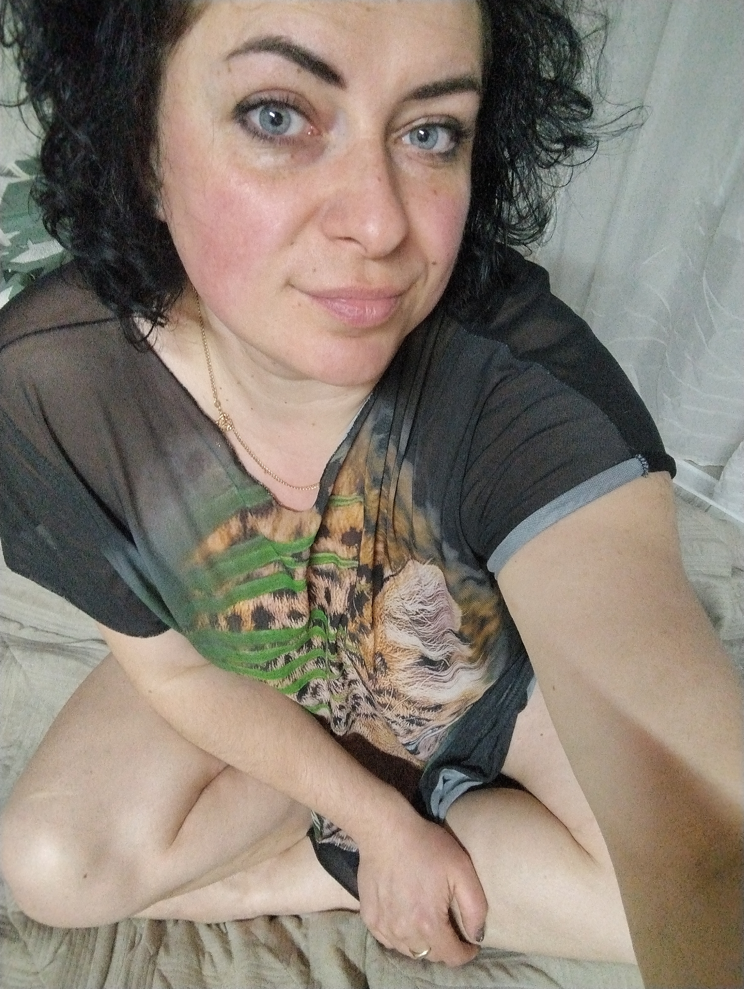 An indoor, eye-level selfie captures a woman with dark, curly hair and striking blue eyes, appearing to be in her 40s or 50s, looking directly at the camera with a soft, gentle smile. She is seated, possibly on a bed or low furniture, with her legs bent or crossed, revealing bare skin.
The woman has a fair complexion with a natural flush on her cheeks. She is wearing a dark, semi-sheer top with short sleeves. The main body of the shirt features a vivid animal print, depicting a tiger or leopard among green foliage. A thin gold chain necklace with a small pendant is visible around her neck, and a simple ring adorns her left ring finger. Her left hand rests on her right thigh.
The background suggests an indoor setting, likely a bedroom or living space in Grishanki, Russia. A textured, light-colored fabric covers the surface she is sitting on, and a sheer, light-colored curtain or drape is visible to her right. The lighting is soft and even, casting no harsh shadows. No text is visible in the image.