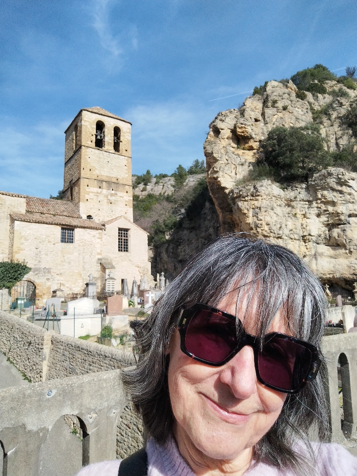 A woman wearing oversized sunglasses stands in front of a weathered stone building with a bell tower and adjacent cemetery. The building appears to be an old church with light-colored stone walls and a tile roof. Behind the church is a steep, rocky cliff with sparse vegetation. The woman is dressed in a light-colored sweater. The sky is clear and blue, and the scene is bathed in sunlight.