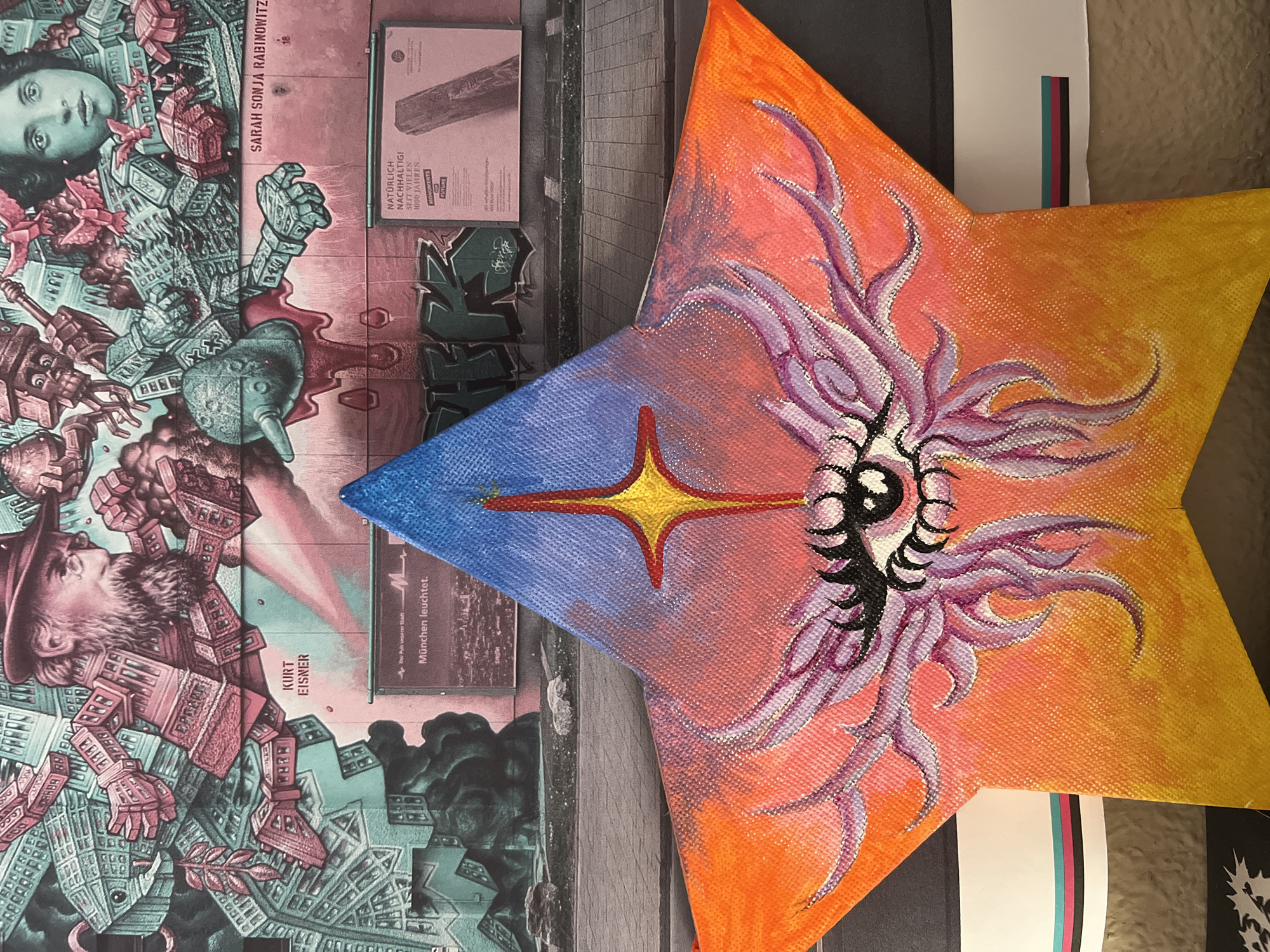 The image shows a close-up, indoor shot featuring a multi-colored, star-shaped canvas leaning against a flat surface covered with printed material. The canvas is divided into color gradients, with a blue section at the top transitioning into orange and then yellow towards the points of the star. A stylized eye is painted on the canvas, surrounded by swirling purple tendrils. A golden star with a red outline is positioned at the center where the blue and orange sections meet.
The surface beneath the canvas is covered with printed materials, including posters or artwork. One section includes text with names "SARAH SONJA RABINOWITZ" and "KURT EISNER." Another section features German text that reads, "NATÜRLICH NACHHALTIG! SEIT VIELEN 1000 JAHREN." Part of a black and white photograph depicting a tall building with a repetitive window pattern is visible in the background.
The context does not seem to imply any scene from the city of Madrid, Spain.