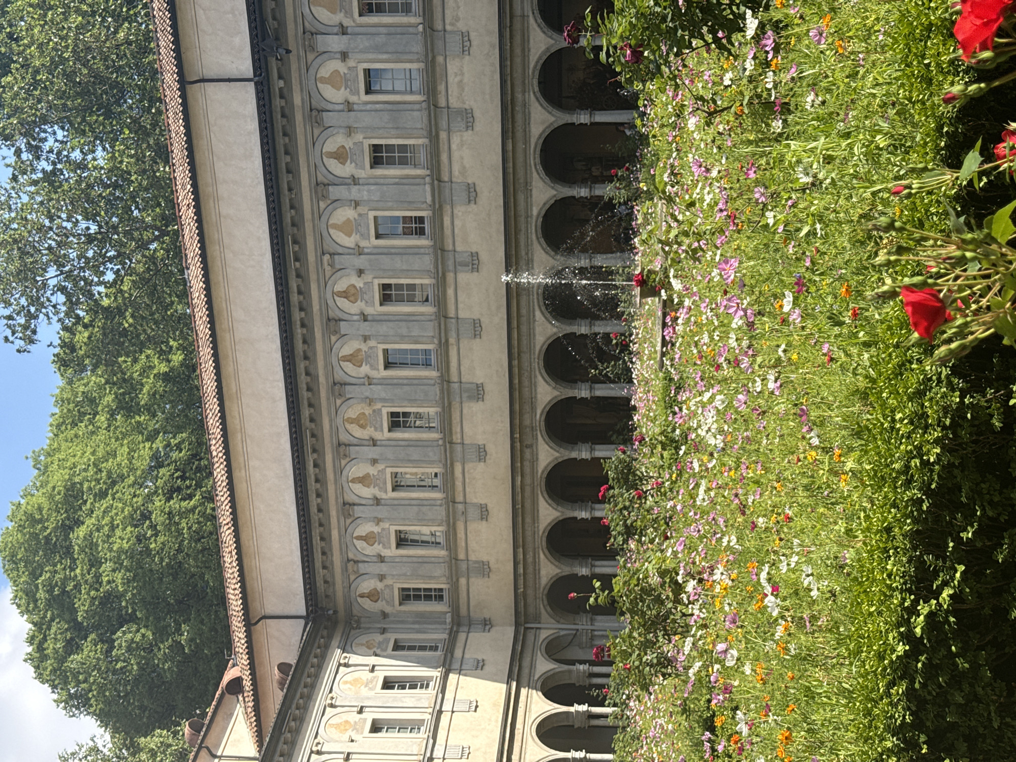 The image displays a serene courtyard scene in what appears to be a historic European setting, consistent with Milan, Italy. A grand, multi-story building occupies the left side of the frame, constructed from a light-colored stone or stucco. Its facade features classical architectural elements, including a series of regularly spaced rectangular windows on the upper floors. Above each window, decorative medallions or busts are integrated into the stonework. The ground floor showcases an elegant arcade of numerous rounded arches, forming a cloister or portico. The building is capped with a traditional reddish-brown tiled roof.
Adjacent to the building, filling the right side of the image, is a vibrant and lush garden or flower bed. It is densely planted with various green foliage and an abundance of small, colorful flowers in hues of white, pink, purple, orange, and yellow, creating a rich and diverse display. A small fountain is active within the garden, with water gracefully spraying upwards, adding movement to the tranquil environment. In the background, on the far left, large, verdant trees with full canopies rise against a clear blue sky, suggesting the extent of the grounds or a park nearby.
The scene is bathed in bright daylight, indicating clear and sunny weather, likely morning or early afternoon. The overall atmosphere is peaceful and beautiful, evoking a sense of calm and historical charm. No people are visible, and there is no discernible text within the image.