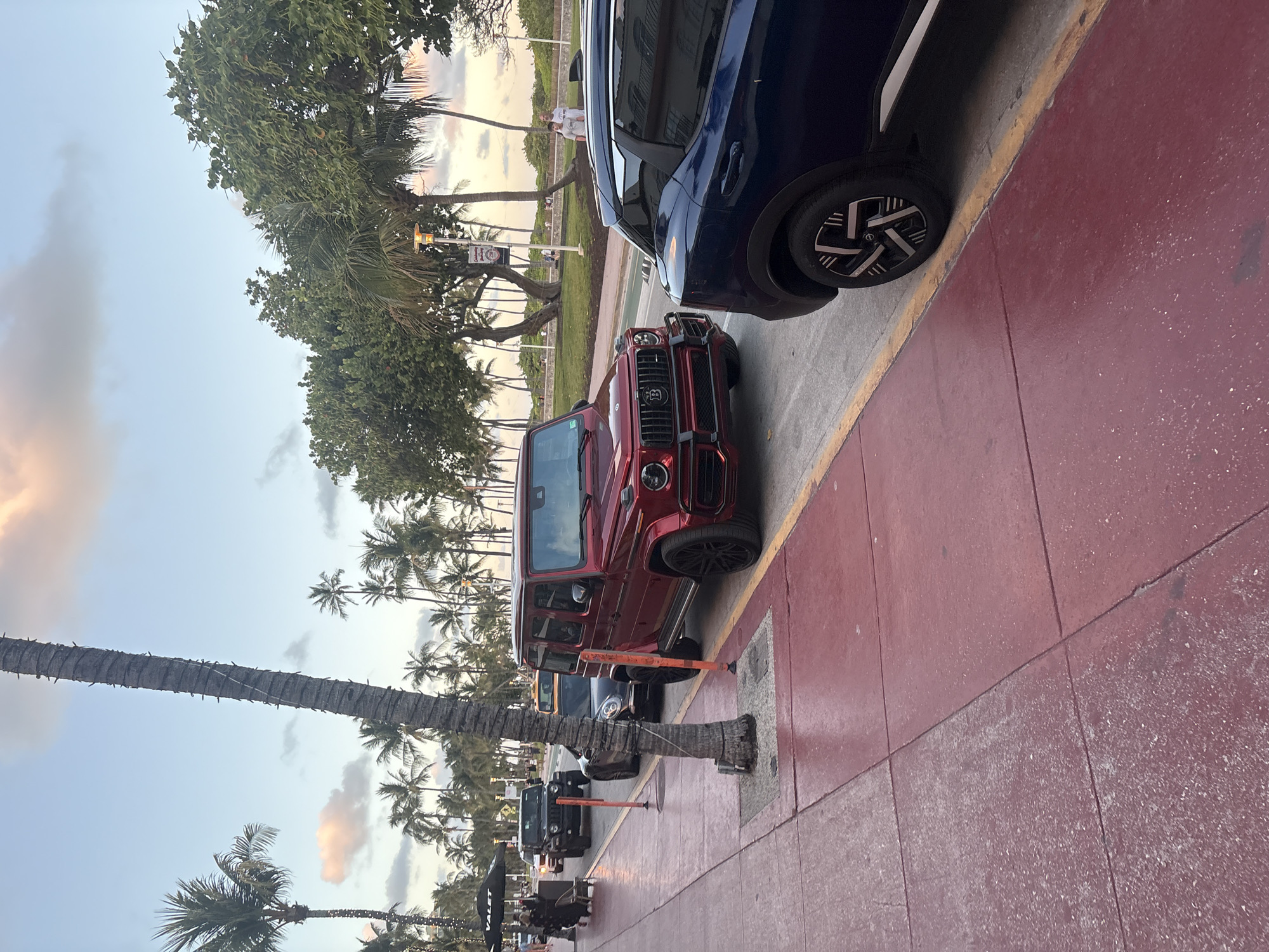 The image captures a street scene, likely at dusk or dawn, given the soft, warm hues in the sky. The perspective is from a low angle, looking across a wide red sidewalk towards a street lined with parked cars and lush tropical trees.
In the foreground, a prominent red Brabus G-Wagen is parked by a yellow curb line on the street. Its distinctive grille with the "B" logo is clearly visible. Behind it, a dark blue SUV is also parked. The sidewalk is a distinct reddish-brown color, showing signs of wear and tear in places.
Further down the street, several other vehicles are parked, including a black SUV. The street is bordered by a green median with numerous palm trees and other leafy green trees, suggesting a tropical or subtropical environment. Some palm trees have string lights wrapped around their trunks. A streetlight and a sign displaying the word "Hotel" are visible amongst the trees.
In the background, on the other side of the street and green median, more palm trees and sparse vegetation line what appears to be a beach or park area. A few people can be seen walking in the distance. To the far left, a tall palm tree with a textured trunk stands prominently, also adorned with lights. Near the lower left, a black "VALET" sign is visible, indicating a service area.
The sky exhibits a gradient of light blue, soft grey, and warm pink/orange clouds, characteristic of sunrise or sunset. The overall atmosphere appears tranquil, capturing a typical evening or morning scene in a resort-like location such as Miami Beach.