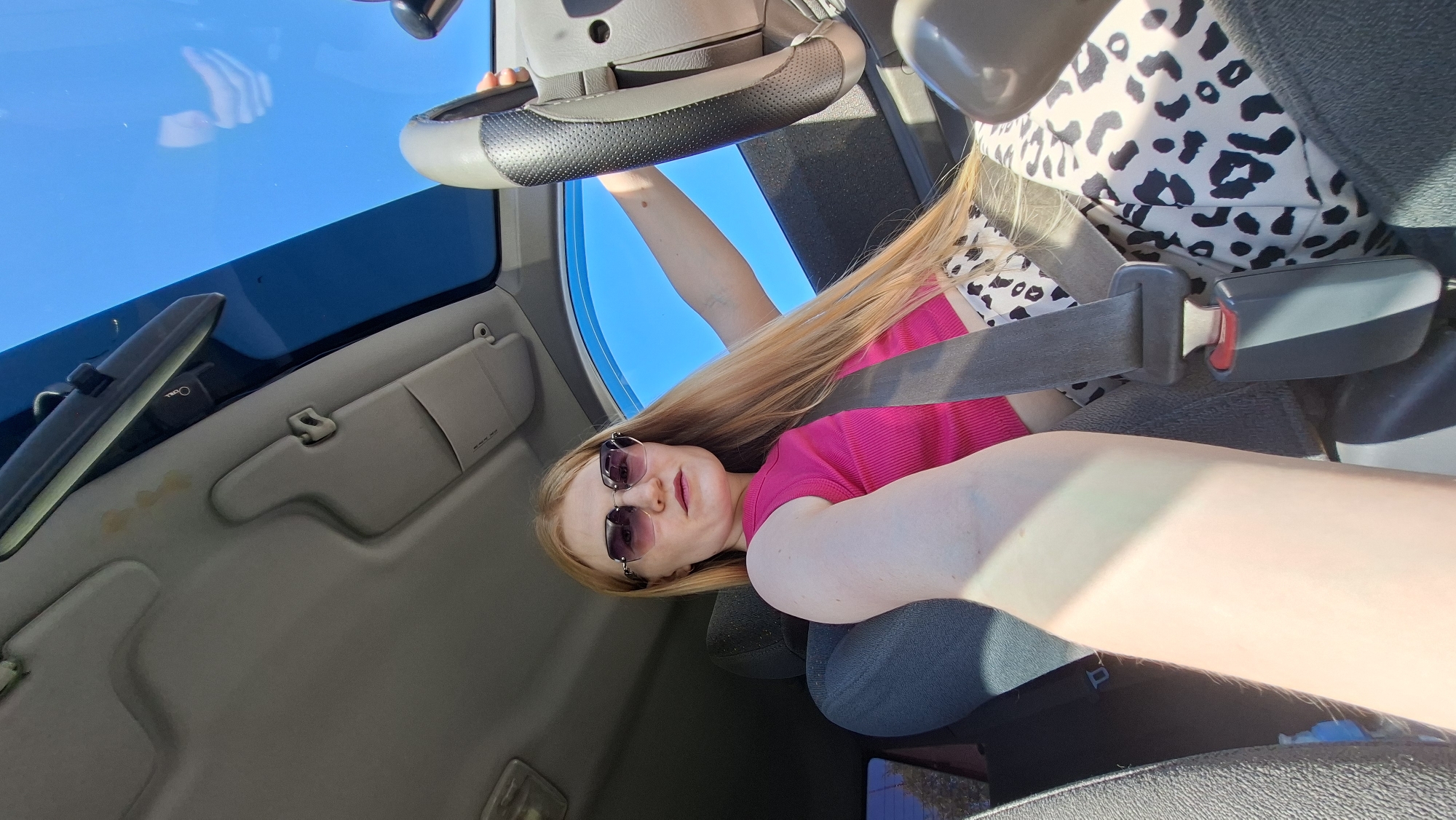 A fair-skinned person with long, light-blonde hair and sunglasses is seated in a car wearing a pink shirt and animal print top, secured with a seatbelt. The image is taken from a low angle inside the vehicle, showcasing the steering wheel, dashboard, and a clear blue sky visible through the windshield. Stains are apparent on the car's interior roof lining. The location in the city of Chaikovsky, Russia, cannot be determined from the image.