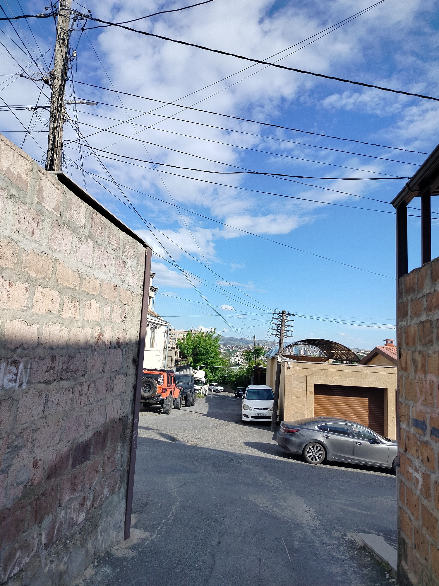 A sunny day in Yerevan, Armenia, reveals a view down a narrow street framed by brick walls. Power lines crisscross the blue sky dotted with clouds. A row of parked cars, including an orange Jeep and a grey Mazda, lines the street. A white van is parked further down. In the distance, a cityscape and distant hills are visible. A utility pole stands prominently near the left brick wall. Buildings are visible beyond the cars and greenery.