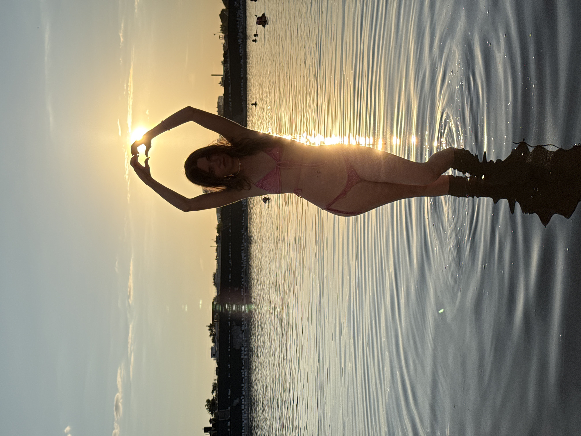 A young woman stands in shallow water, her arms raised above her head, forming a heart shape with her hands around the setting sun. She is wearing a pink bikini. The water is calm with visible ripples, and her reflection is visible below her. In the background, there is a distant shoreline with structures and trees, and other people are visible in the water. The sky is a gradient of orange and blue, indicating sunset. The scene is likely set in Sol-Iletsk, Russia, known for its salt lakes.