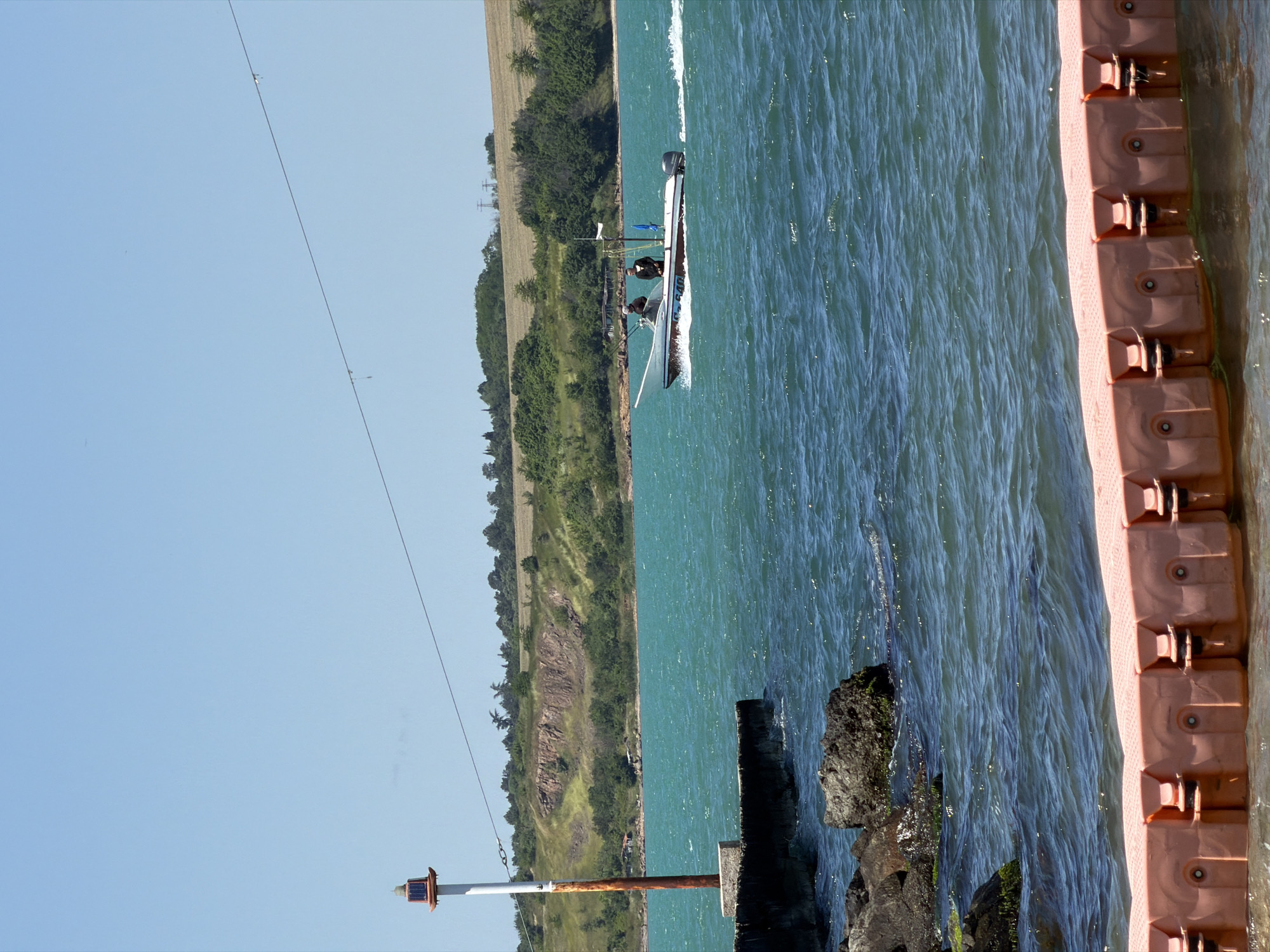 The image depicts a stretch of water, likely the Black Sea, next to Burgas, Bulgaria, with an orange pontoon dock on the right side and the coastline on the left. A motorboat with two people aboard is moving in the water. A rusty metal pole with a red and white lamp is visible in the bottom left corner, and a power line stretches across the top of the frame. The sky is clear and blue. Rocks covered in moss can be seen in the water.