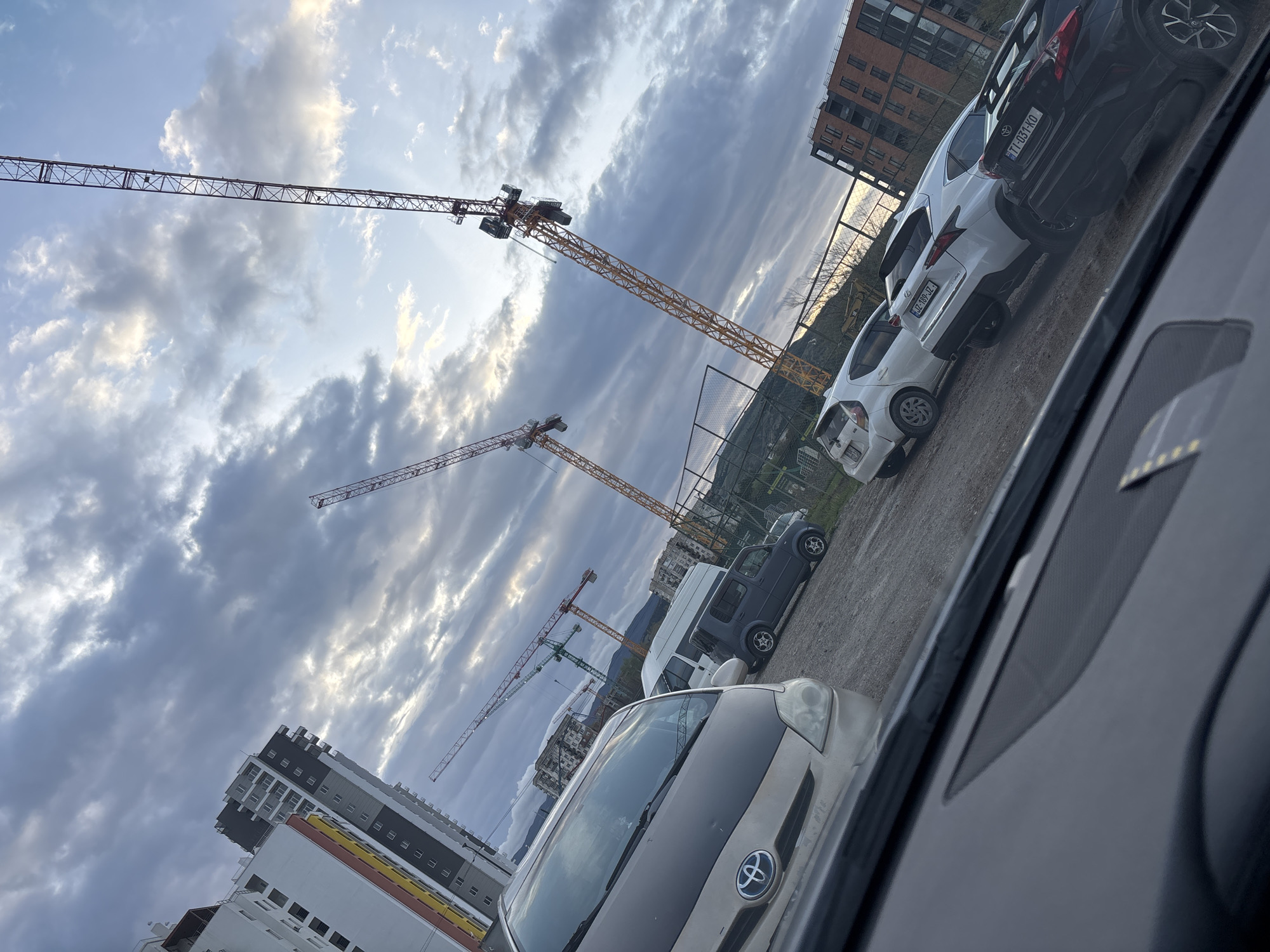 The image, taken from inside a car, shows a view of construction cranes against a cloudy sky in Tbilisi, Georgia. The photo is taken at an angle, skewing the perspective. In the foreground, parts of the car interior are visible. A silver car with a Toyota emblem is partially in the shot. Behind it, several parked vehicles are lined up on a gravel surface, including white and black sedans. In the background, a chain-link fence separates the parking area from a slightly raised terrain. Construction cranes of various sizes dominate the skyline, contrasting with the partially visible buildings. The cloudy sky is illuminated with patches of sunlight. The setting appears to be a construction site or adjacent area within Tbilisi, Georgia.