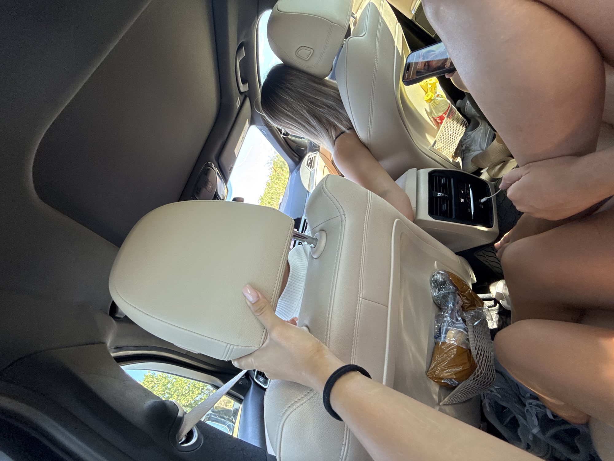 The image shows the interior of a car, likely a BMW given the design of the headrests and air vents. The viewpoint is from the back seat, looking forward. Several people are present, though not all are fully visible. In the foreground, a person's legs are visible, suggesting they are sitting in the back. To the left, another person's arm is reaching up, holding onto the headrest of the front seat. Their hand, with manicured nails, is positioned near the top of the headrest. A black hair tie is visible on their wrist.
In the middle ground, a woman with blonde hair is seated in the passenger seat, facing away from the camera. She appears to be leaning her head back against the headrest. Her back and shoulders are visible, and she is wearing a dark-colored top.
On the back of the front passenger seat, a mesh pocket contains a plastic-wrapped package of food, possibly snacks, and some other items. In the footwell of the back seat, a pair of light-colored sneakers are visible, along with a grey blanket or clothing item.
The car's interior is light-colored leather. Sunlight streams in from the windows, illuminating the scene. The exterior view through the windows shows trees and a bright sky, suggesting it is daytime. A smartphone is visible on the seat to the right, displaying a person's face on the screen. The air vents in the back of the center console are also visible, with a temperature reading of 19.5 displayed. The image was taken in the city of Lehancea, Romania.