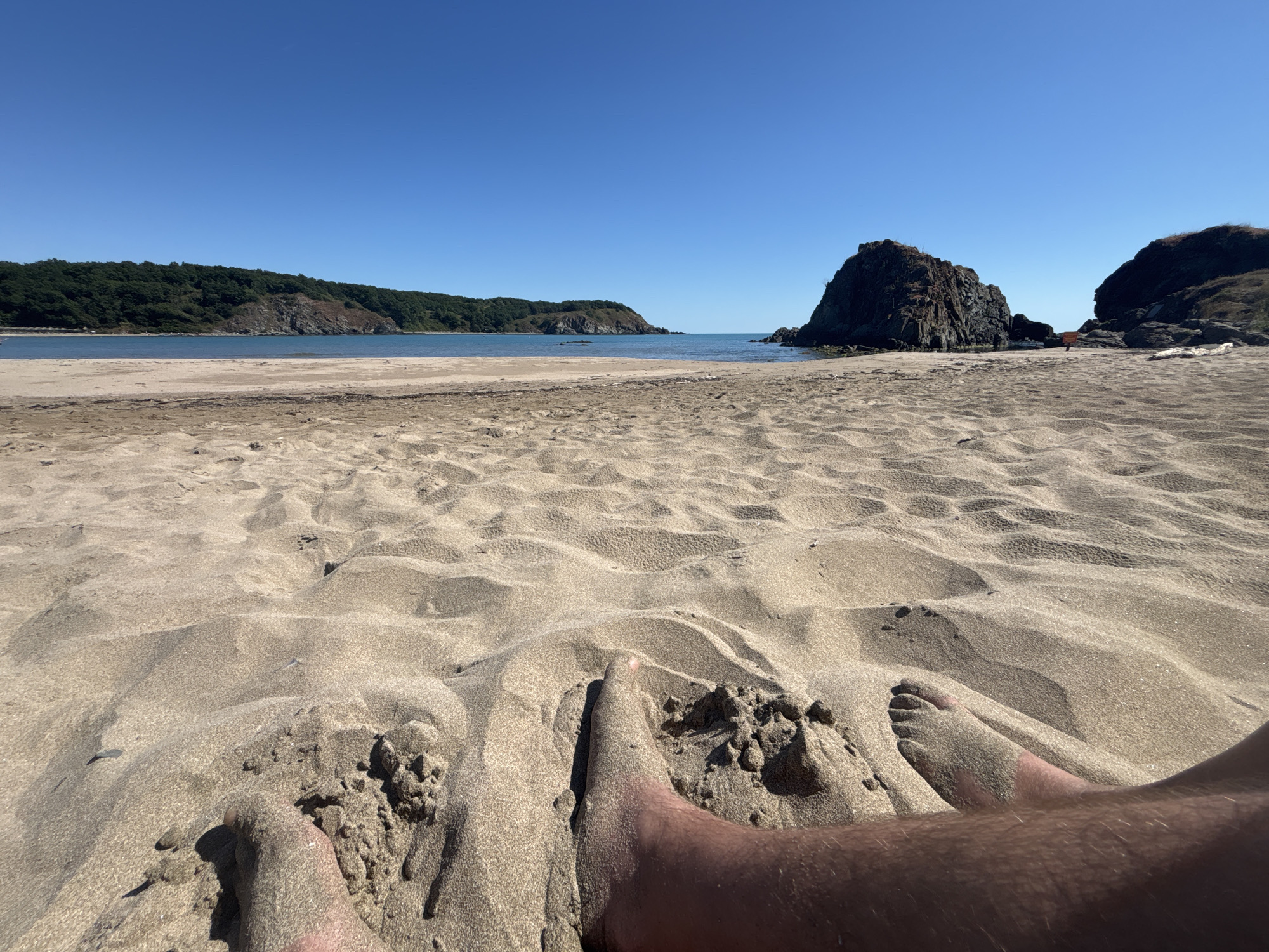In the city of Rezovo, Bulgaria, a first-person perspective shot captures a sandy beach under a clear blue sky. The foreground is dominated by sand dunes, with a person's bare feet partially submerged and covered in sand. The middle ground reveals a tranquil sea and a sandy shoreline, leading to a rugged, rocky outcropping jutting out into the water. In the background, a verdant, tree-covered hill slopes down towards the sea, forming a picturesque coastal landscape.