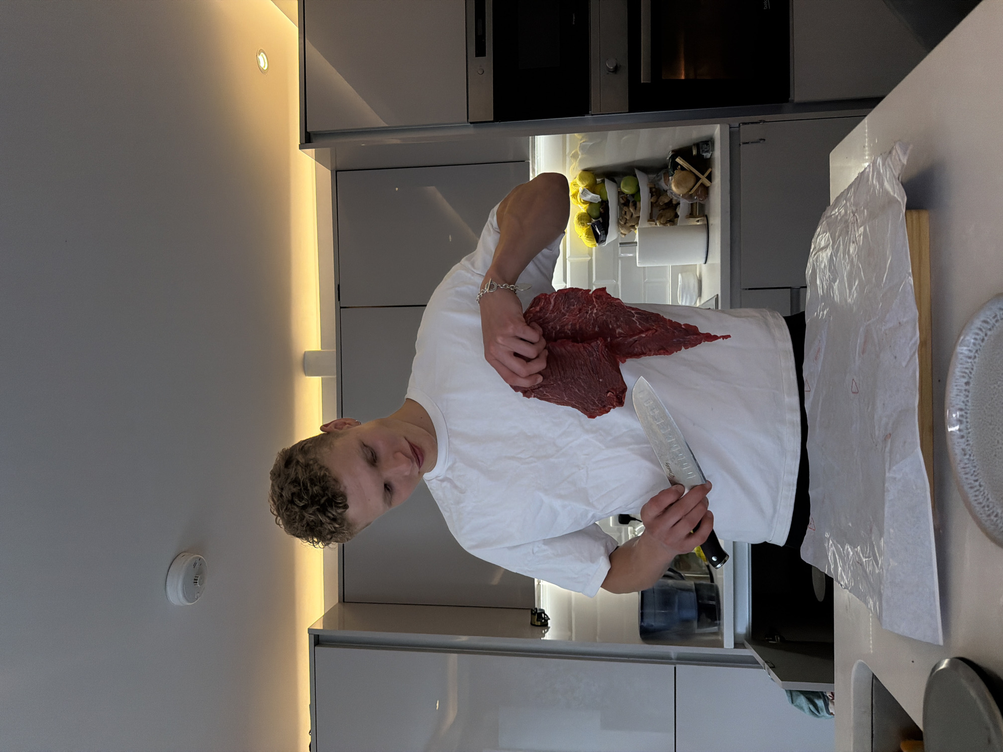 The image features a young man in a modern kitchen, likely in London, United Kingdom. He has light skin, short curly blonde hair, and is dressed in a white t-shirt, wearing a silver chain bracelet on his left wrist. He is holding a large, raw piece of red meat, possibly flank steak, in his left hand and a substantial chef's knife in his right. He looks directly at the camera with a slight, friendly smile, appearing ready to begin cutting or preparing the meat.
The contemporary kitchen setting includes sleek light grey cabinets and light-colored countertops. Integrated appliances, including what appears to be an oven or microwave, are visible in the background. Warm LED strip lighting illuminates the wall behind the man and underneath the upper cabinets, contributing to a clean and inviting ambiance. A white smoke detector is mounted on the ceiling to the left. On the countertop to the right, there's a cutting board covered with white parchment paper or plastic wrap, next to a textured white plate. Inside an open cabinet or refrigeration unit, clear containers hold fresh produce like lemons and ginger, alongside a roll of paper towels. The blade of the chef's knife clearly shows the text "Stainless Steel" and a partial brand name, possibly "Qucheur".