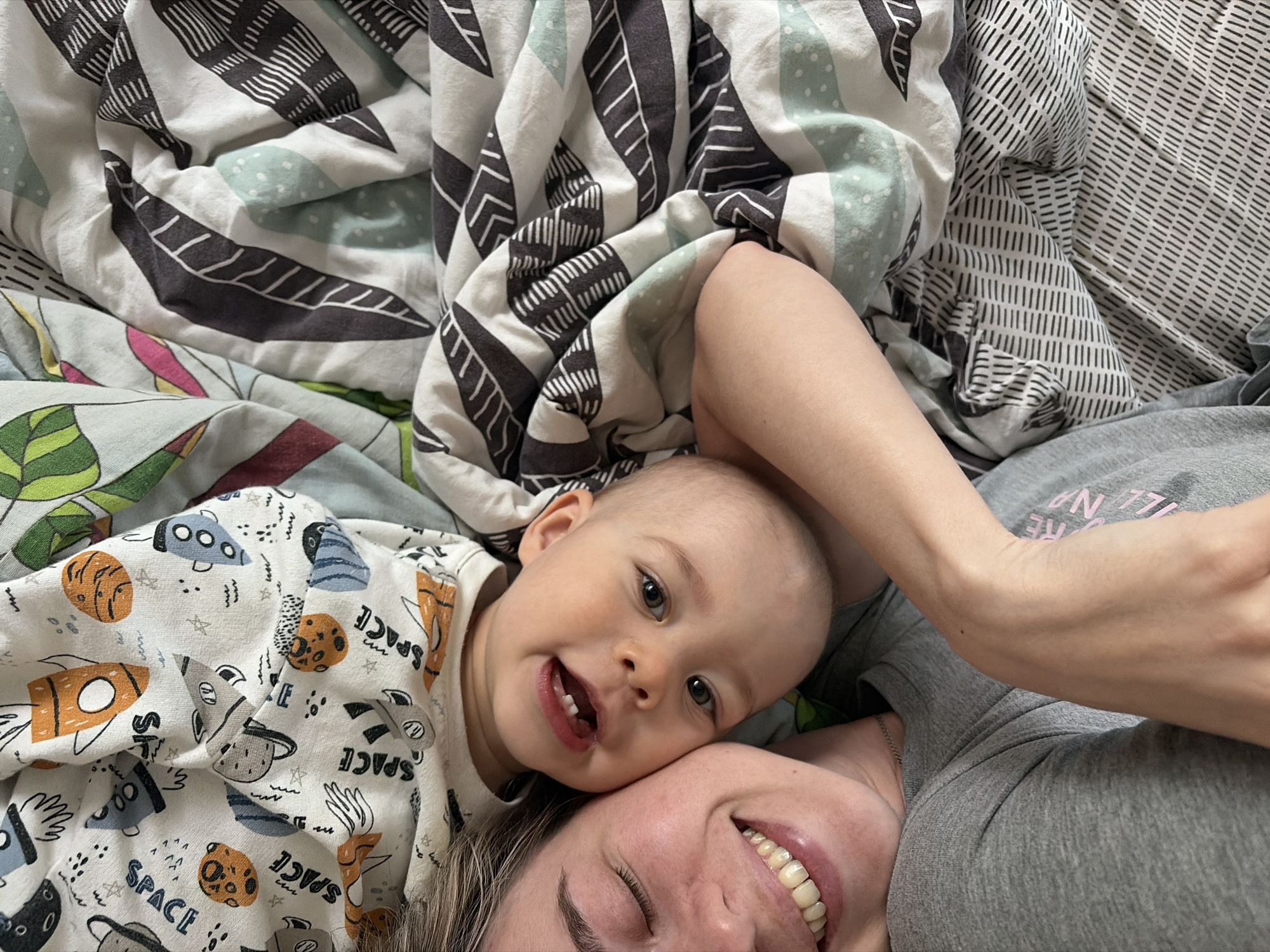 A photograph taken in what is likely an indoor setting shows a woman and a baby lying down, presumably together on a bed. The baby, wearing a white shirt with a pattern of spaceships and planets, is looking directly at the camera with a smile. The woman is smiling with her eyes closed and the baby is lying down next to her. There is a colorful blanket with patterns of leaves, and abstract shapes.
Based on the location, this image was likely taken in the city of Chaikovsky, Russia.