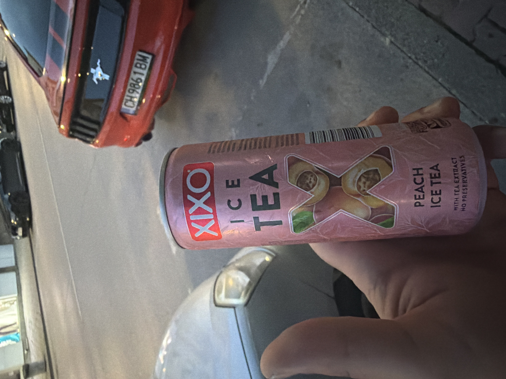 The image, rotated 90 degrees clockwise, shows a person's hand holding a can of iced tea in an outdoor setting. The can is primarily pink and white, labeled "XIXO ICE TEA" with "PEACH ICE TEA" prominently displayed, accompanied by an illustration of peaches and green leaves. Additional text on the can specifies "WITH TEA EXTRACT" and "NO PRESERVATIVES." A barcode is visible at the top.
In the background, a red car is parked, its rear visible. It features two black stripes running down the trunk and a chrome emblem of a galloping horse, indicative of a Ford Mustang. A Swiss license plate, "CH 9861 BM," is clearly attached to the car, indicating its registration in Switzerland. This contradicts the provided location context of Shumen, Bulgaria. The car is positioned on asphalt or concrete pavement. Partially visible around the red car are other vehicles, suggesting a street or parking area. The lighting suggests it is daytime, possibly late afternoon, with diffused light and soft shadows. No specific weather conditions are evident. The scene depicts a casual moment, with no discernible activity beyond holding the beverage.