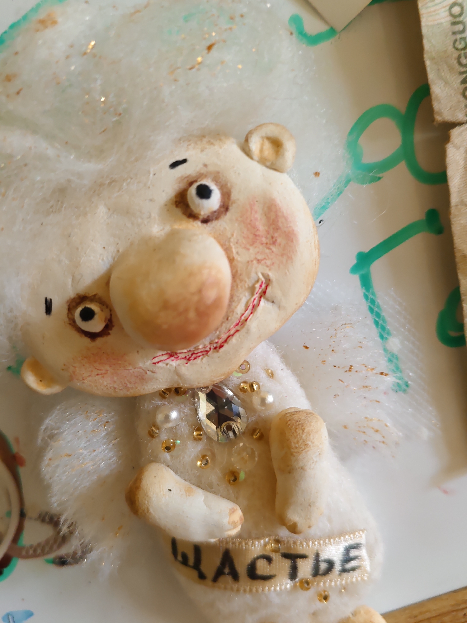 The image shows a close-up of a handmade plush toy, possibly a "good luck charm" or a decorative figurine. The toy is made of light-colored material, likely felt or soft fabric, and has a beige-colored face with a large, round nose. Its eyes are black dots, and it has rosy cheeks with faint red blush marks. The toy has a stitched red mouth that appears to be smiling. It is adorned with various small, shiny embellishments, including pearls and gold beads, as well as a clear, faceted gemstone on its chest. A ribbon with the Cyrillic word "СЧАСТЬЕ" (which translates to "happiness" in English) is attached to the toy's lower body. The background is out of focus but appears to be a flat surface with some green markings and a portion of what looks like a banknote, suggesting it might be a display or a craft project. The toy is in the foreground and is the primary subject. There are no people visible in the image, and no specific location in Rostov-on-Don, Russia is depicted.