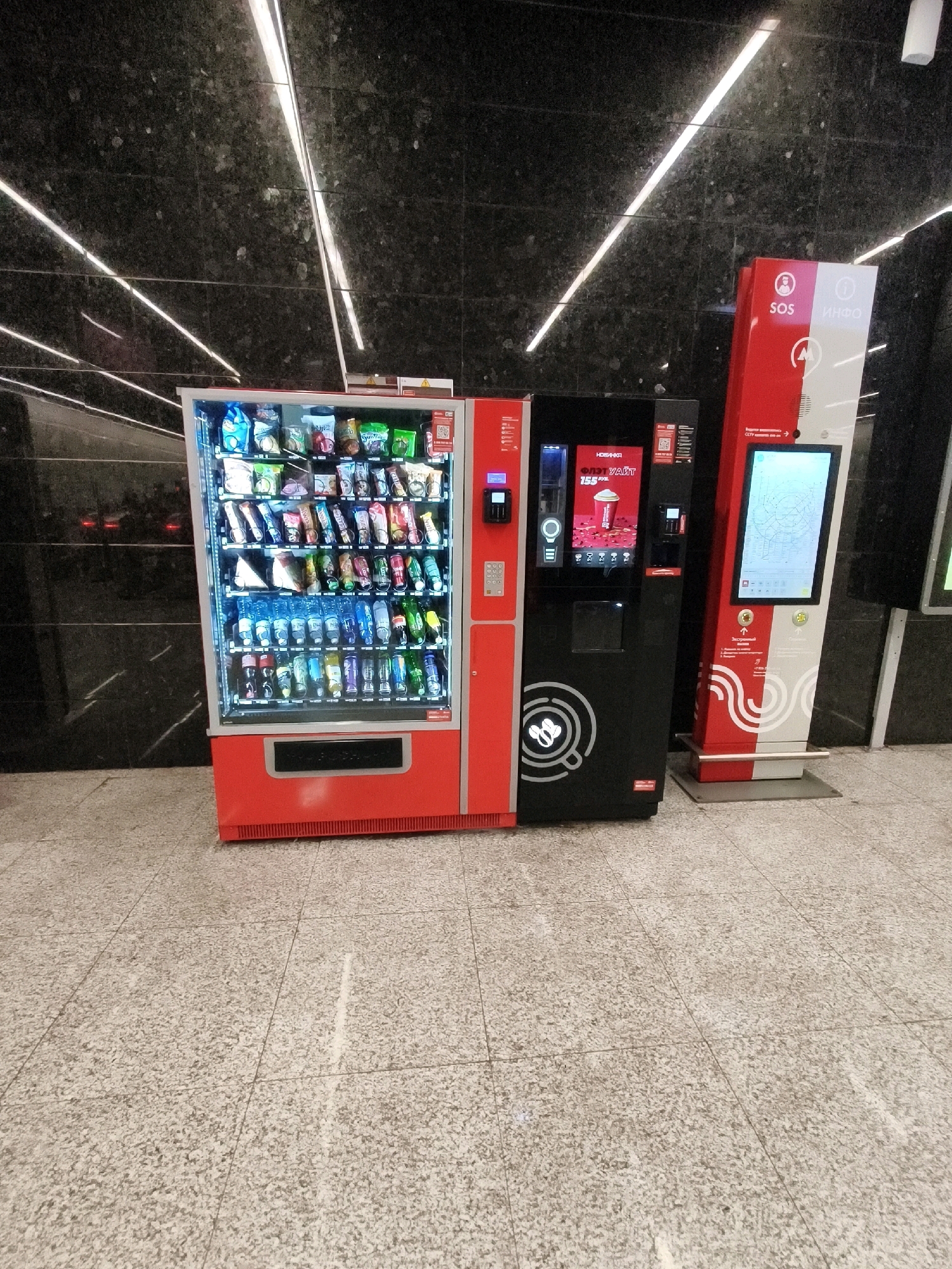 In an indoor environment with a dark, speckled wall and a tiled floor, three vending machines are visible. The leftmost machine is red and filled with various snacks and drinks. To its right is a black machine with a screen displaying a coffee cup graphic and text, and a coin/bill slot. Further to the right is a tall, red and white machine with a large screen showing a subway map. The lighting in the scene is provided by overhead fluorescent lights. The machines are positioned side-by-side in what appears to be a public area.