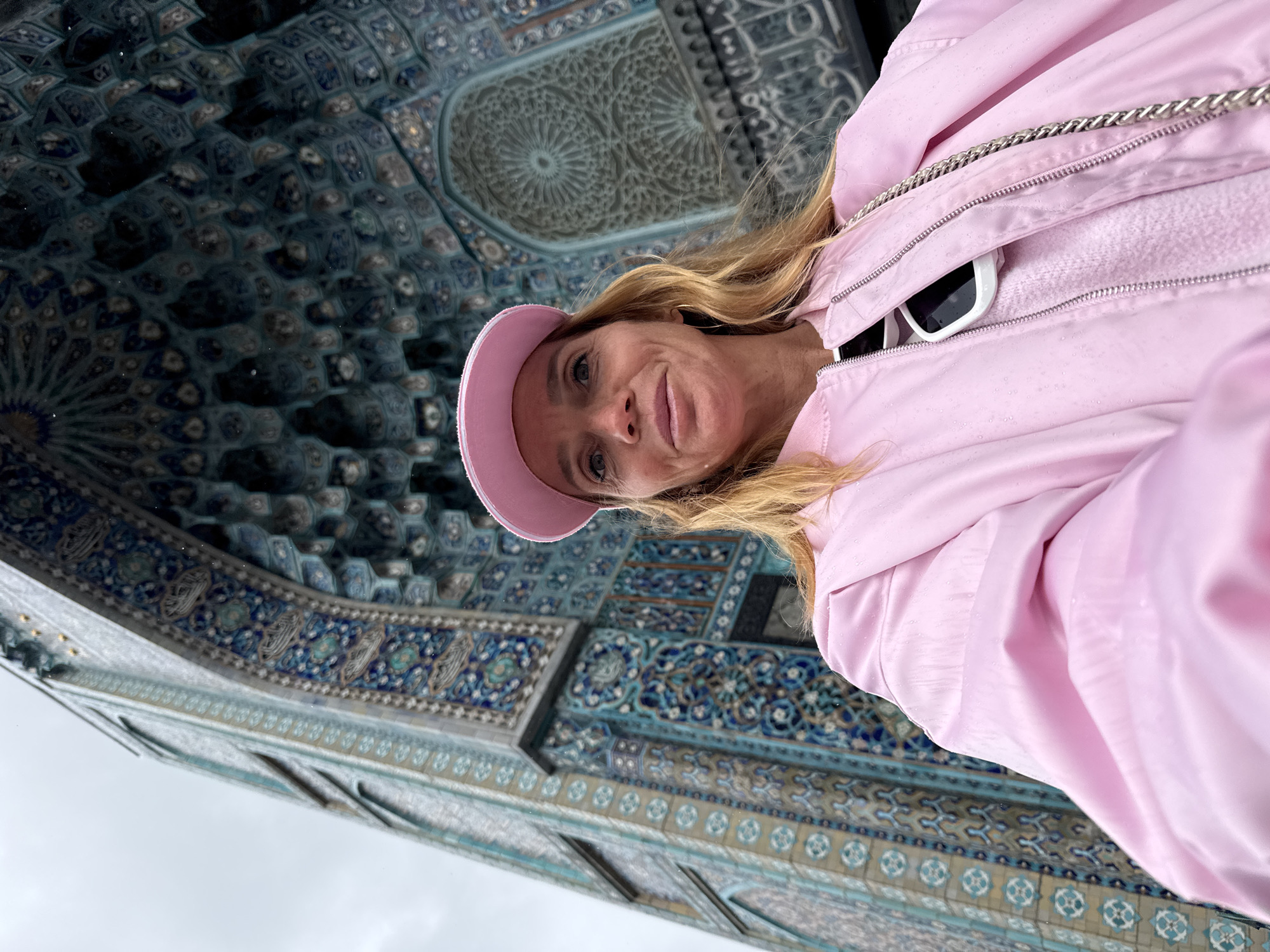 A low-angle, rotated, eye-level shot shows a woman standing beneath the exterior facade of a building, which is ornate with blue and grey mosaic tile. The woman has blond hair and is wearing a pink baseball cap and a light pink zip-up jacket. Sunglasses are visible in the open jacket. The building is ornate with mosaic tiles and carvings. The weather appears overcast. The building is likely in Saint Petersburg, Russia.