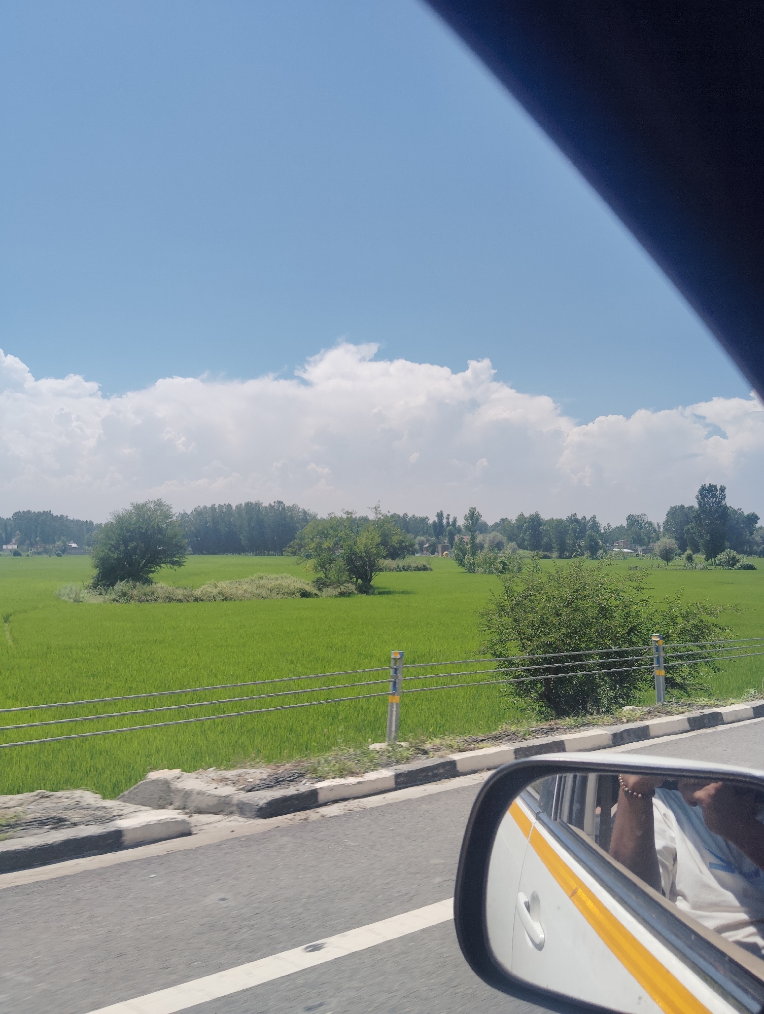 The image is a photograph taken from inside a vehicle, looking out at a rural landscape in Awantipora, India. The foreground features the side mirror of the vehicle, reflecting the interior and a person's arm with a bracelet. The vehicle's yellow and white stripe is visible on the car's body. Beyond the vehicle, a road with a white line marking runs parallel to a fence made of wire and reflective posts. A vibrant green paddy field stretches across the mid-ground, bordered by a line of trees in the distance. The sky above is a bright blue, dotted with fluffy white clouds.