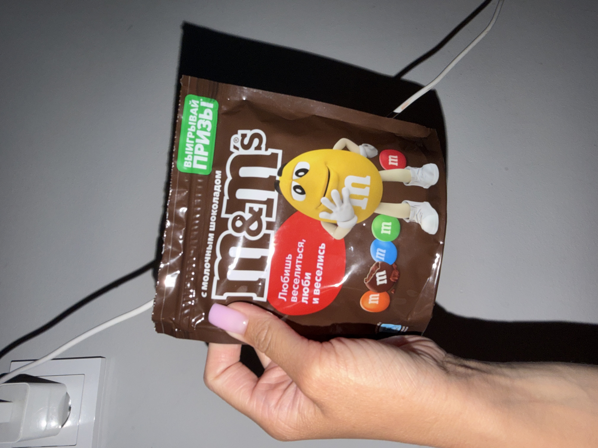 A hand with light pink fingernail polish is holding a brown M&M's candy bag. The bag is partially unzipped, revealing some of the candy inside. The bag has the M&M's logo and a cartoon yellow M&M character. There is text on the bag in Russian, including "С МОЛОЧНЫМ ШОКОЛАДОМ" (with milk chocolate) and a green rectangle with "ВЫИГРЫВАЙ ПРИЗЫ" (Win Prizes). The background is a plain gray wall. To the left of the bag, a white electrical cord is plugged into a white wall outlet. The image appears to be taken indoors, possibly in a residential setting in the city of Sirius, Russia.