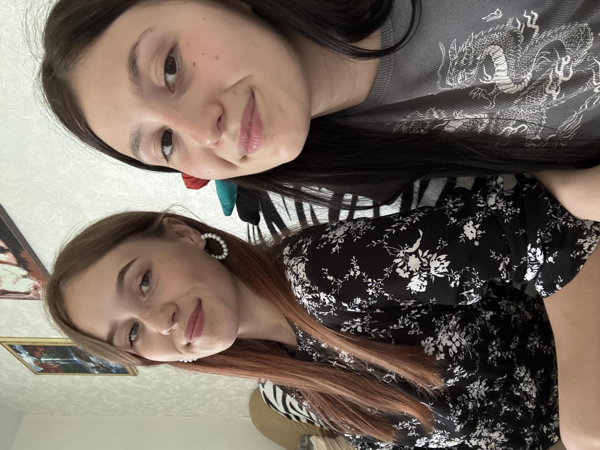 Two young women are posing closely together for a selfie, possibly indoors. The woman at the top has dark hair and is wearing a gray shirt with a dragon design. The woman at the bottom has light brown or red hair, wears pearl earrings, and a black dress with white floral patterns. In the background, there's a painting framed on the wall and what appears to be a zebra print item behind them. The location is not explicitly mentioned or visibly discernable from the image to be Фоки, Russia.
