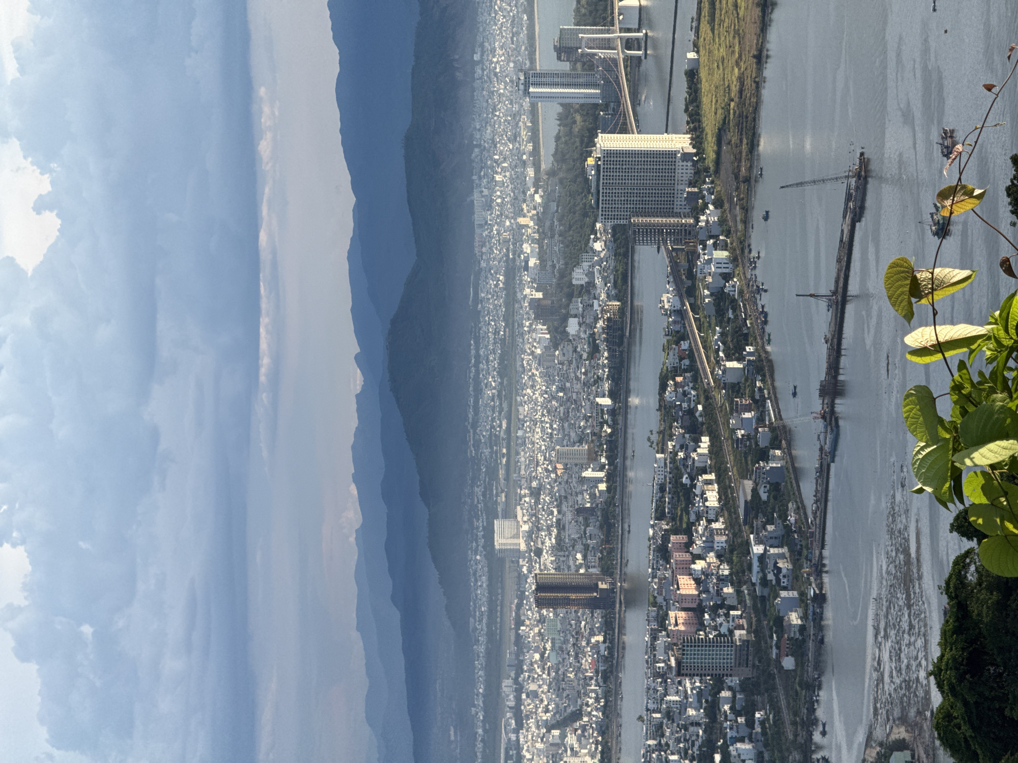 An aerial, wide-angle, horizontal medium shot captures the cityscape of Da Nang, Vietnam, with a river bisecting the frame. In the foreground, lush green foliage partially obscures the view from the top left corner and along the bottom of the frame. The river, a wide expanse of calm, grey water, dominates the lower half of the image. Several small boats are visible on the river, including a cluster of moored vessels and others further out. A long, narrow construction pier extends into the river from the right side of the frame, featuring two prominent cranes and other construction equipment.
Beyond the river, the city unfolds in a dense arrangement of buildings. Numerous mid-rise residential buildings, many with light-colored facades, fill the middle ground. A few taller, modern skyscrapers stand out, including a distinctive dark-colored twin-towered building and a lighter-colored building with a bridge connecting it to the shore. Two bridges span the river, with one notably large, modern bridge with white suspension cables and white pylons visible in the mid-right of the frame. The city stretches towards a hazy horizon, where a dense urban landscape merges with rolling, blue-hued mountains that recede in layers towards the background. The sky above is a mix of soft, grey clouds with patches of bright light, suggesting either early morning or late afternoon. The overall scene depicts a bustling urban environment integrated with its natural surroundings.