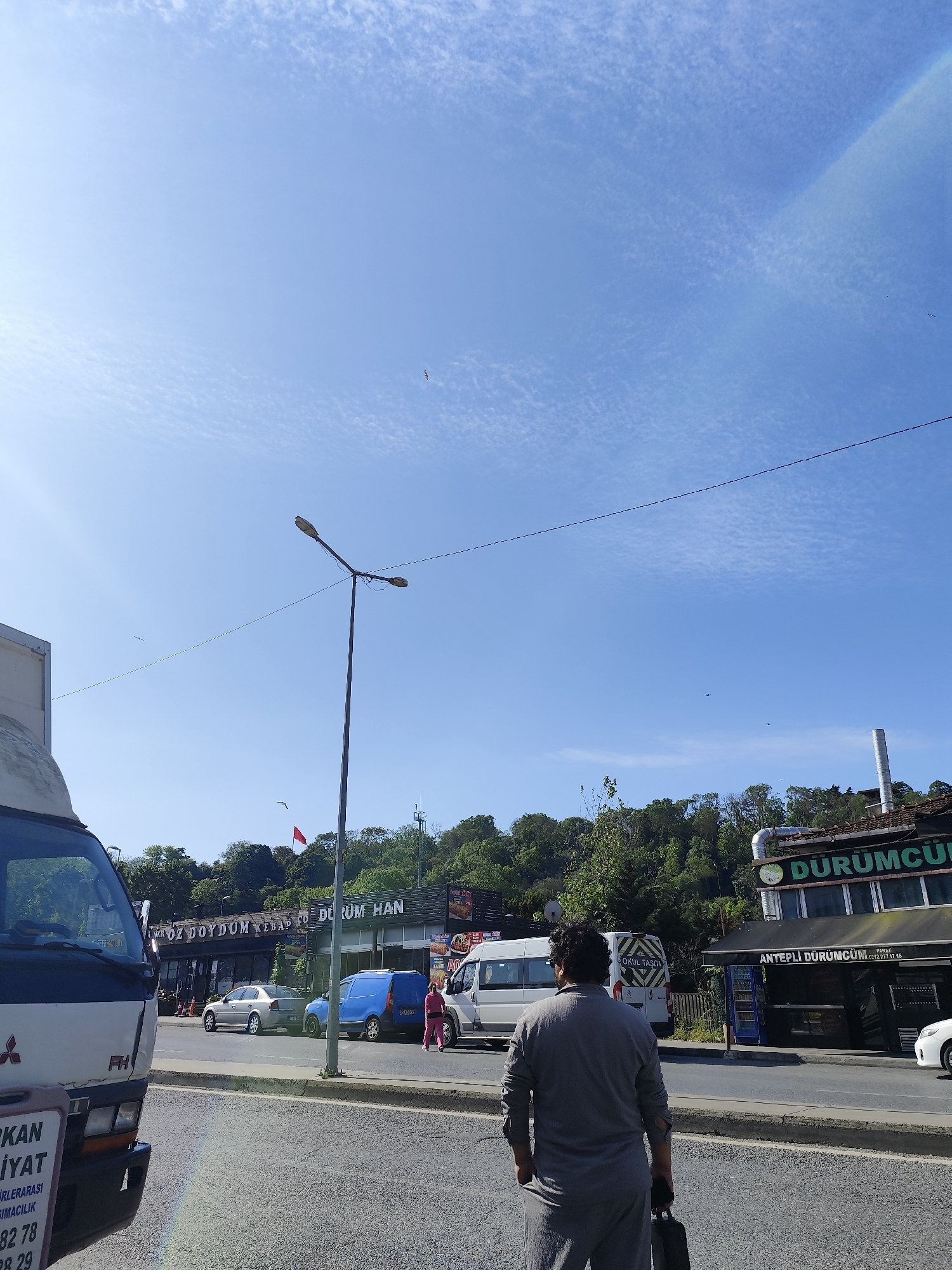 The image depicts an outdoor scene in the Sarıyer district of Istanbul, Turkey, characterized by a blue sky with faint cirrus clouds, indicating clear weather. A man is seen from the back, wearing a gray, full-sleeved outfit, and carrying a black object in his left hand. He is standing on a paved road, possibly a pedestrian crossing or waiting to cross.
Several commercial establishments line the opposite side of the road, including "ÖZ DOYDUM KEBAP CO. DURUM HAN" and "ANTEPLİ DÜRÜMCÜM," both suggesting Turkish cuisine, specifically kebabs and durum. Various vehicles are parked or passing by, including a white van with "OKUL TAŞITI" (School Shuttle) written on it and a white truck with "FH" visible on the front. A utility pole with overhead wires is positioned nearby. The surrounding environment includes green trees and vegetation visible in the background, indicating a wooded or park-like area. A Turkish flag is visible in the mid-ground. The overall scene appears to capture daily life in a commercial and residential area of Sarıyer.