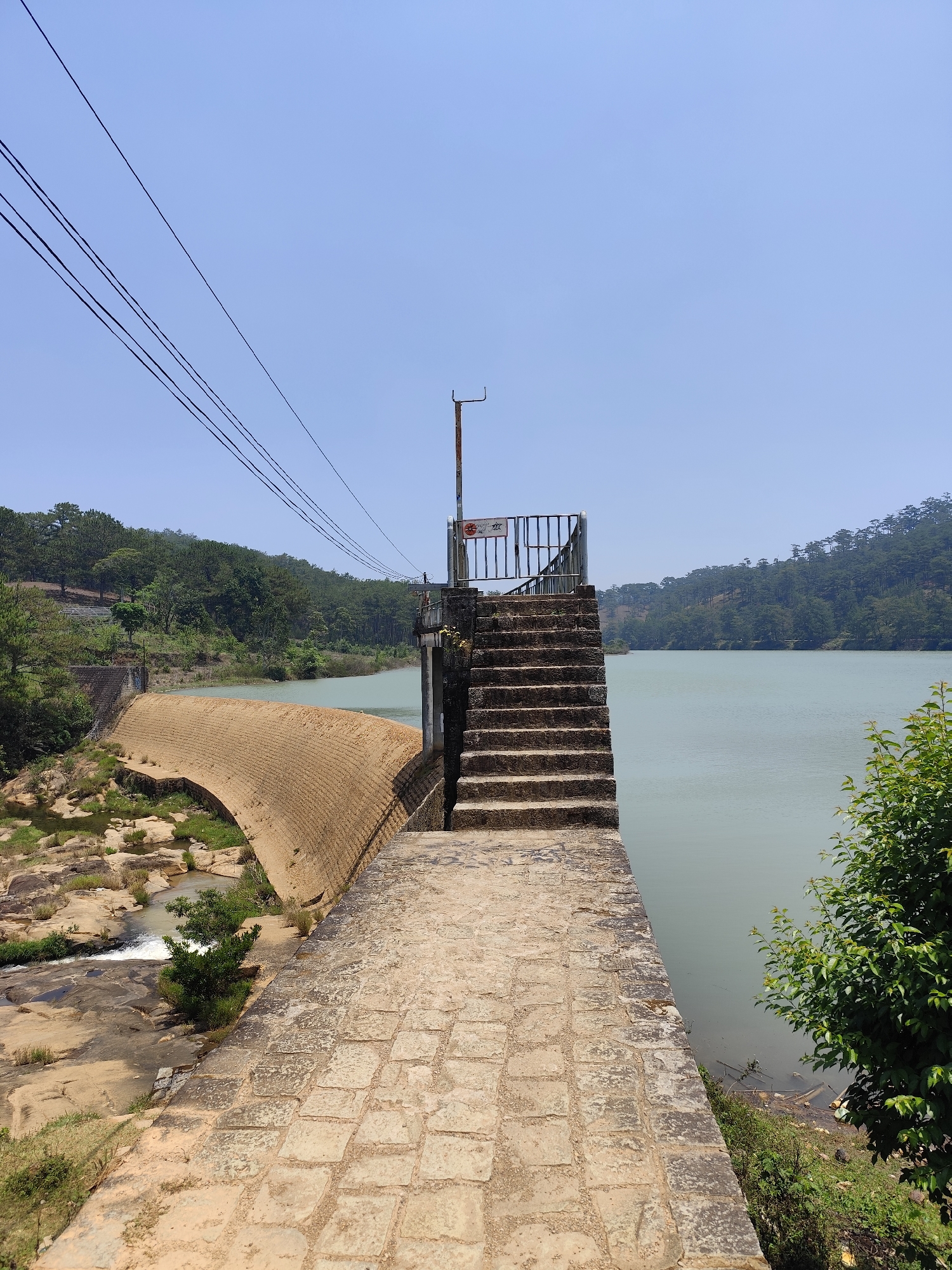 The image depicts a concrete dam and spillway in Lang Biang - Da Lat, Vietnam. The structure has a staircase leading to a small observation platform with metal railings, adjacent to a reservoir. A series of power lines extends across the top left of the image. A curved brick spillway directs water towards a rocky riverbed. The reservoir's surface is calm. Forested hills line the horizon. Vegetation surrounds the dam. The sky is clear and blue.